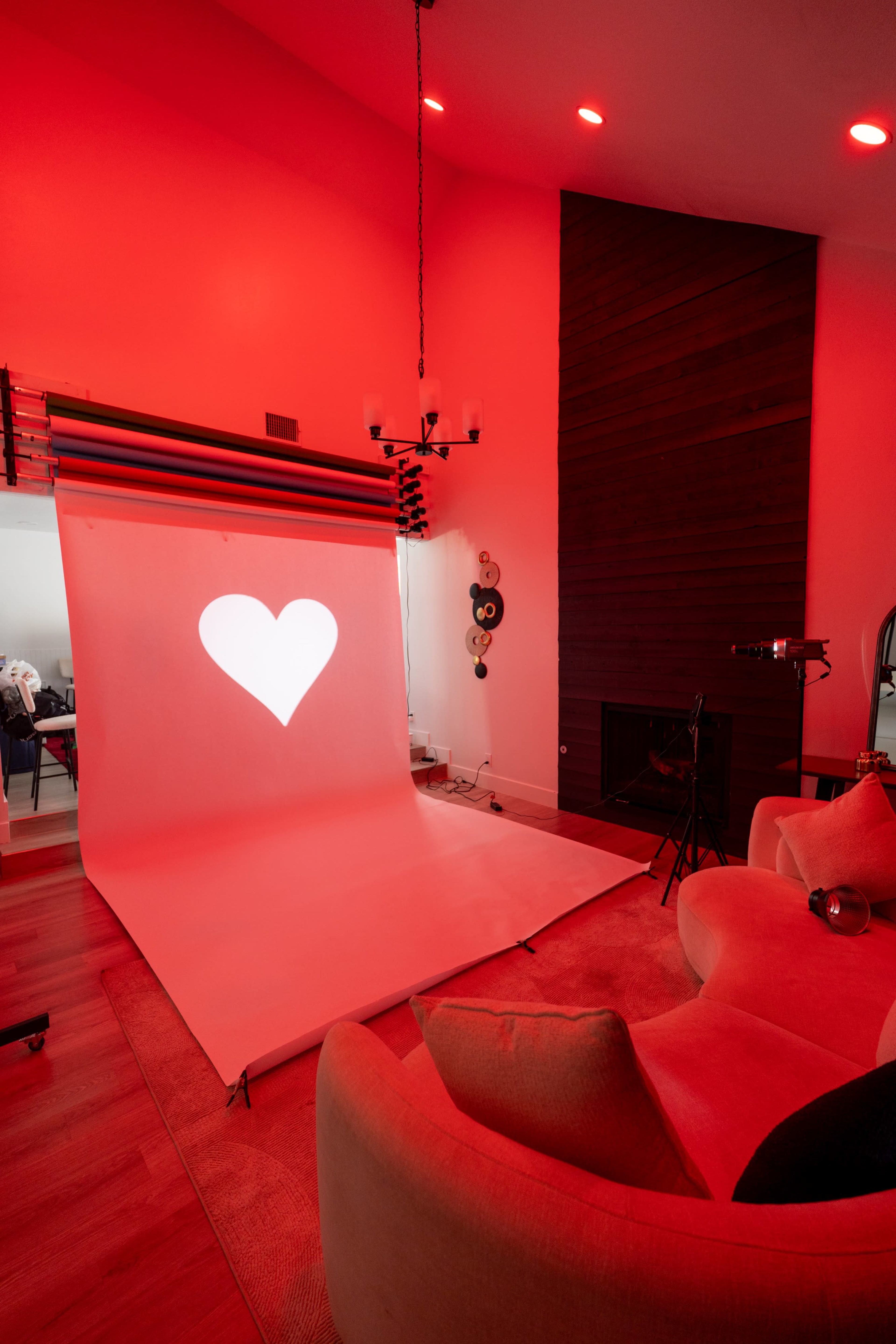A bright red-lit room features a white backdrop with a heart shape projected on it, alongside a cozy seating area.