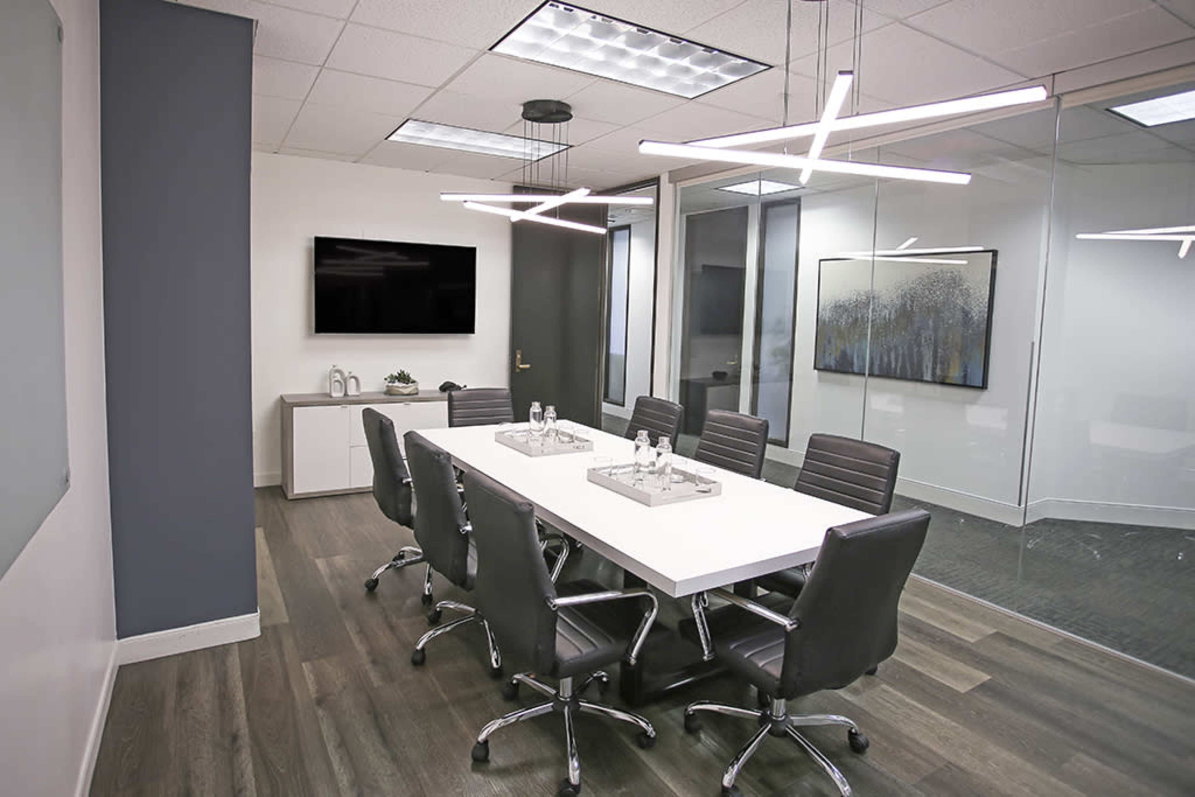 A modern conference room featuring a long white table surrounded by black chairs, with a television mounted on the wall and a glass partition.