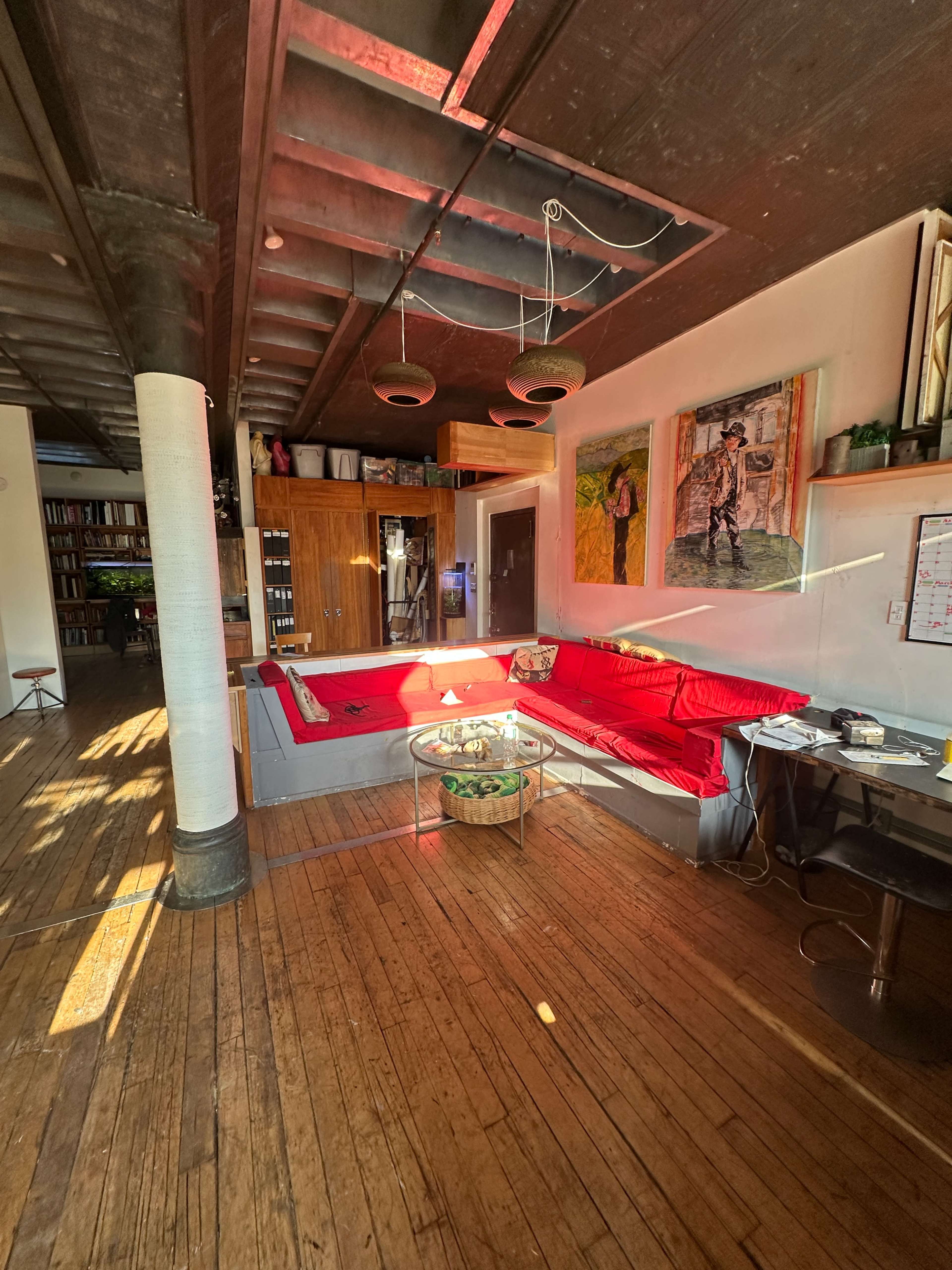 The image shows a spacious living room with hardwood floors, two red sofas, a glass coffee table, and walls adorned with artwork.