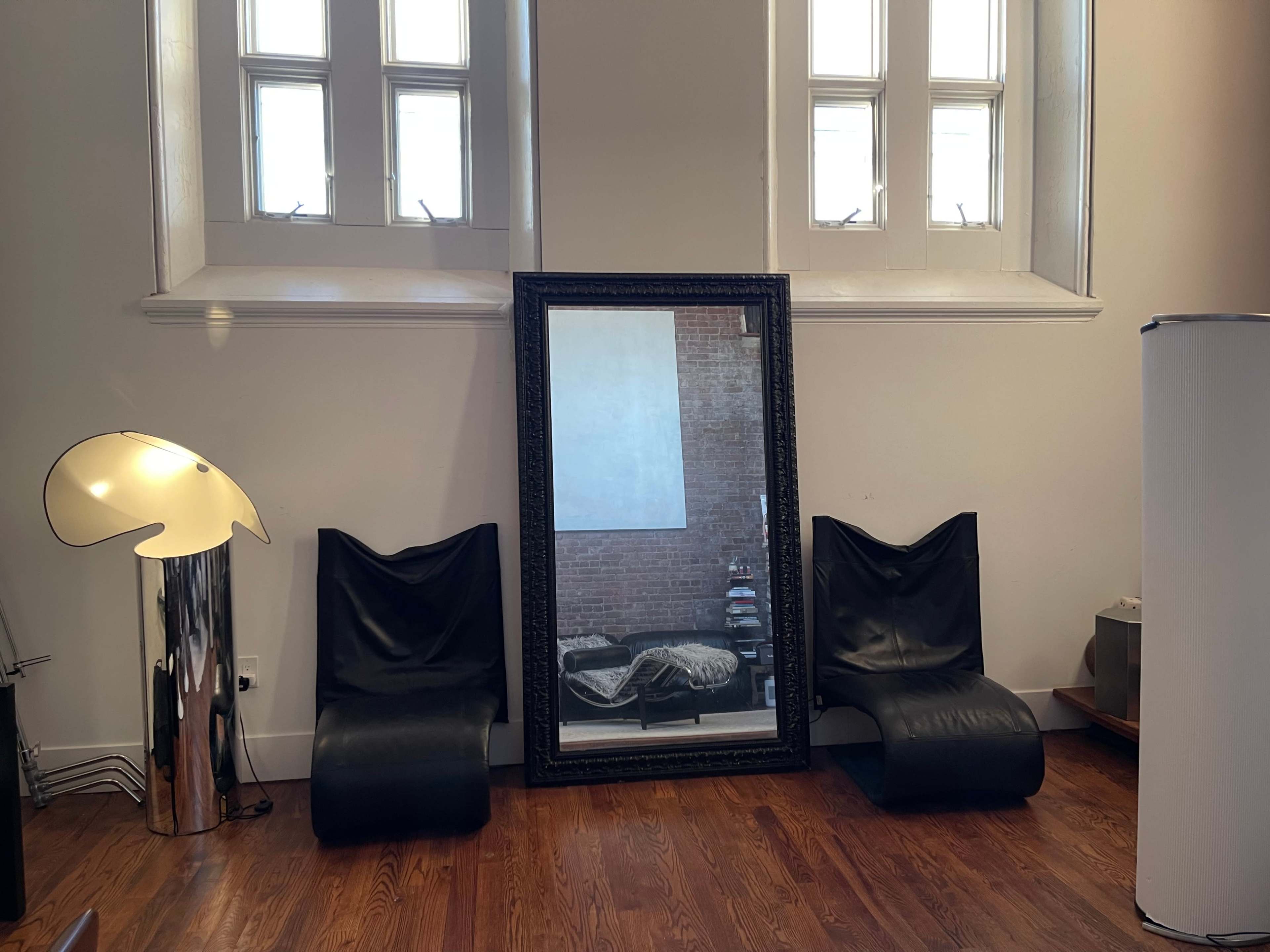 A large ornate mirror leaning against a wall, flanked by two black lounge chairs and a modern floor lamp in a room with wooden flooring and a brick wall.