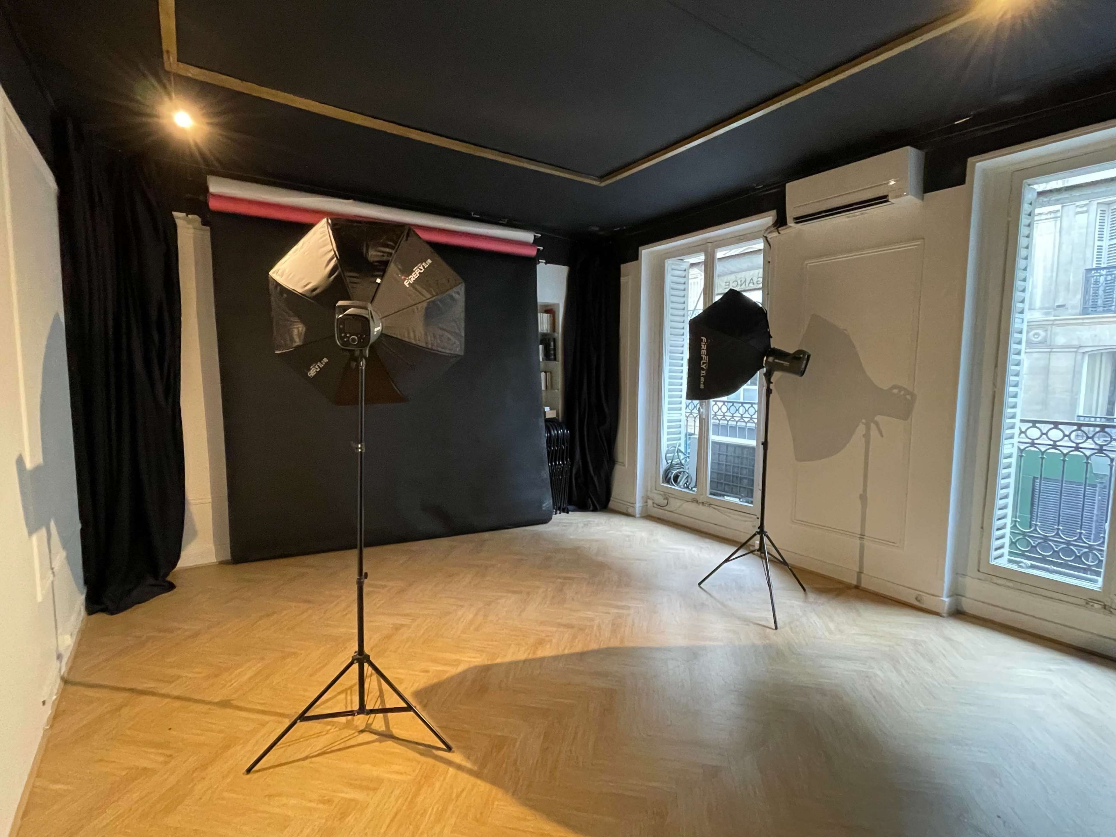 A photo studio featuring two softbox light stands, a black backdrop, and large windows with balcony views.