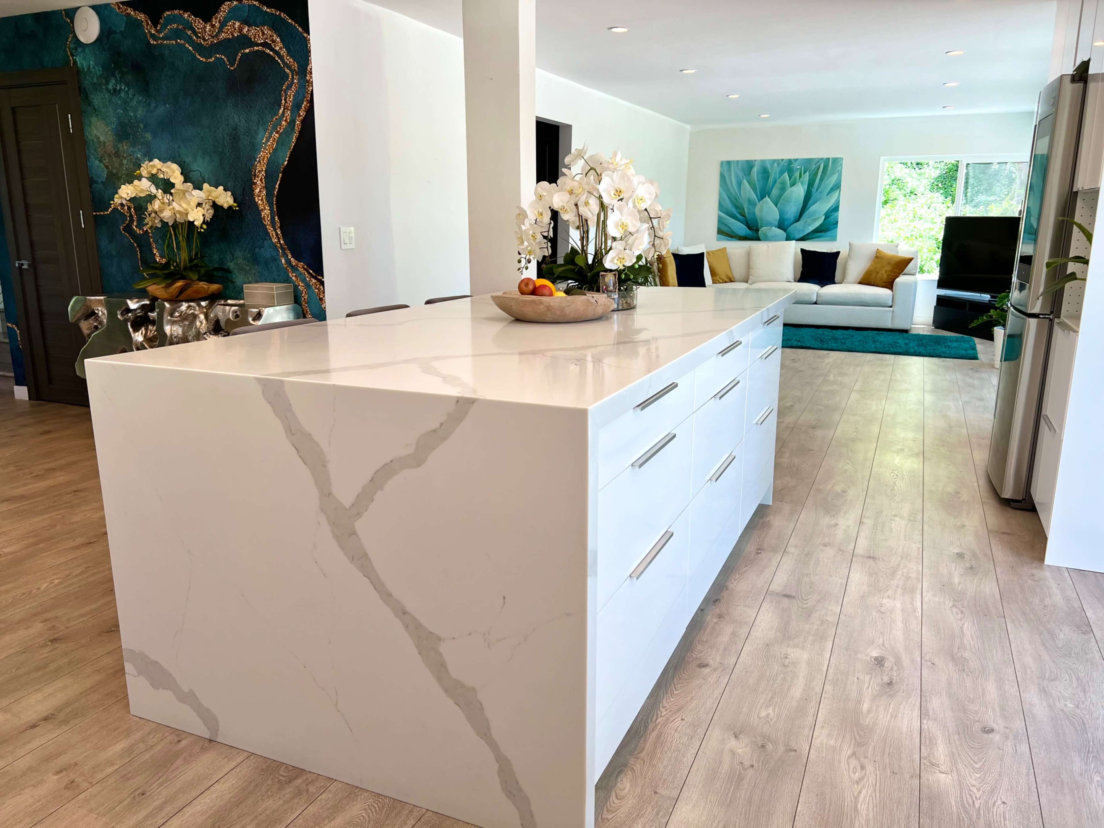 The image shows a modern kitchen featuring a large white marble island with drawers, surrounded by a living area with a turquoise rug and a floral painting on the wall.