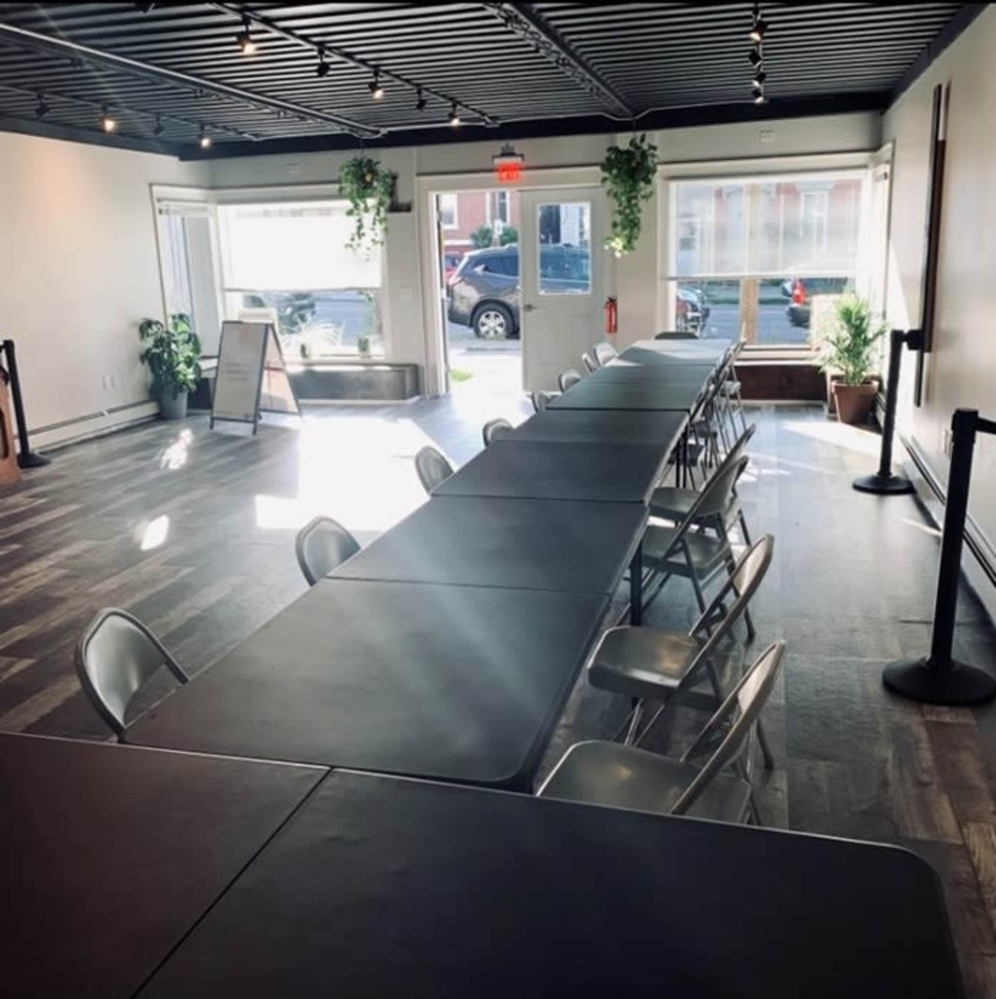 A spacious, well-lit room with a long table and chairs arranged for a meeting or event, with large windows and plants along the walls.