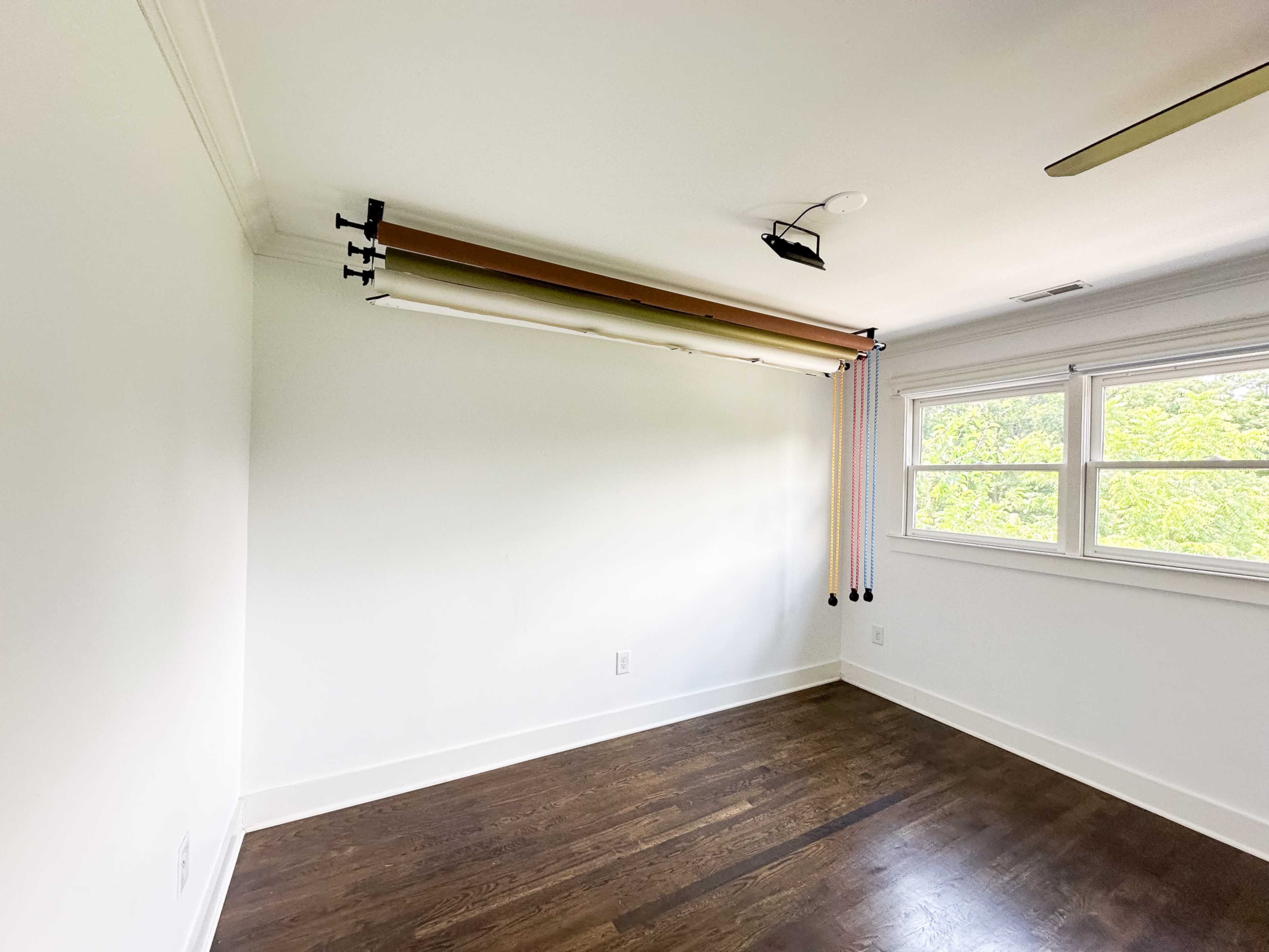 The image shows a mostly empty room with hardwood floors, a large window, and a backdrop setup consisting of rolled fabric suspended from a track along the wall.