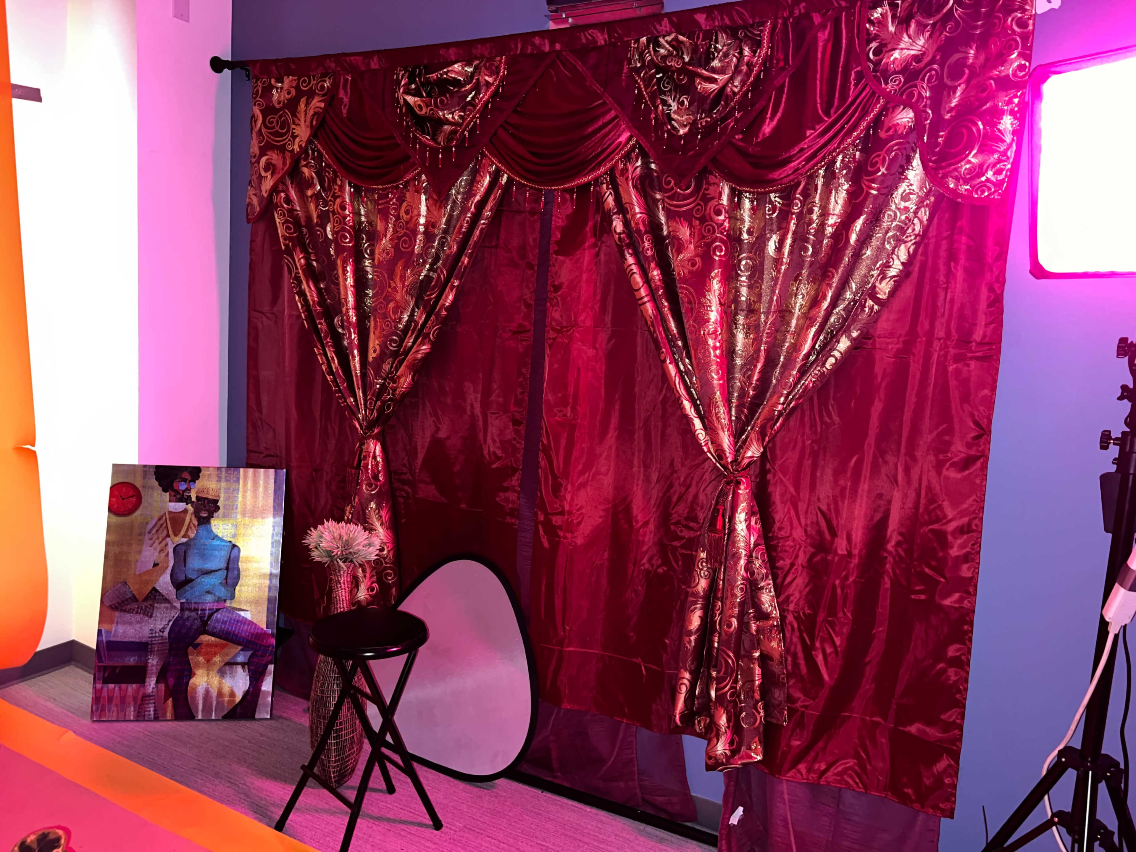 The scene features a backdrop of red curtains with ornate patterns, a framed artwork on the floor, a stool, and a reflector disk in a brightly lit room.