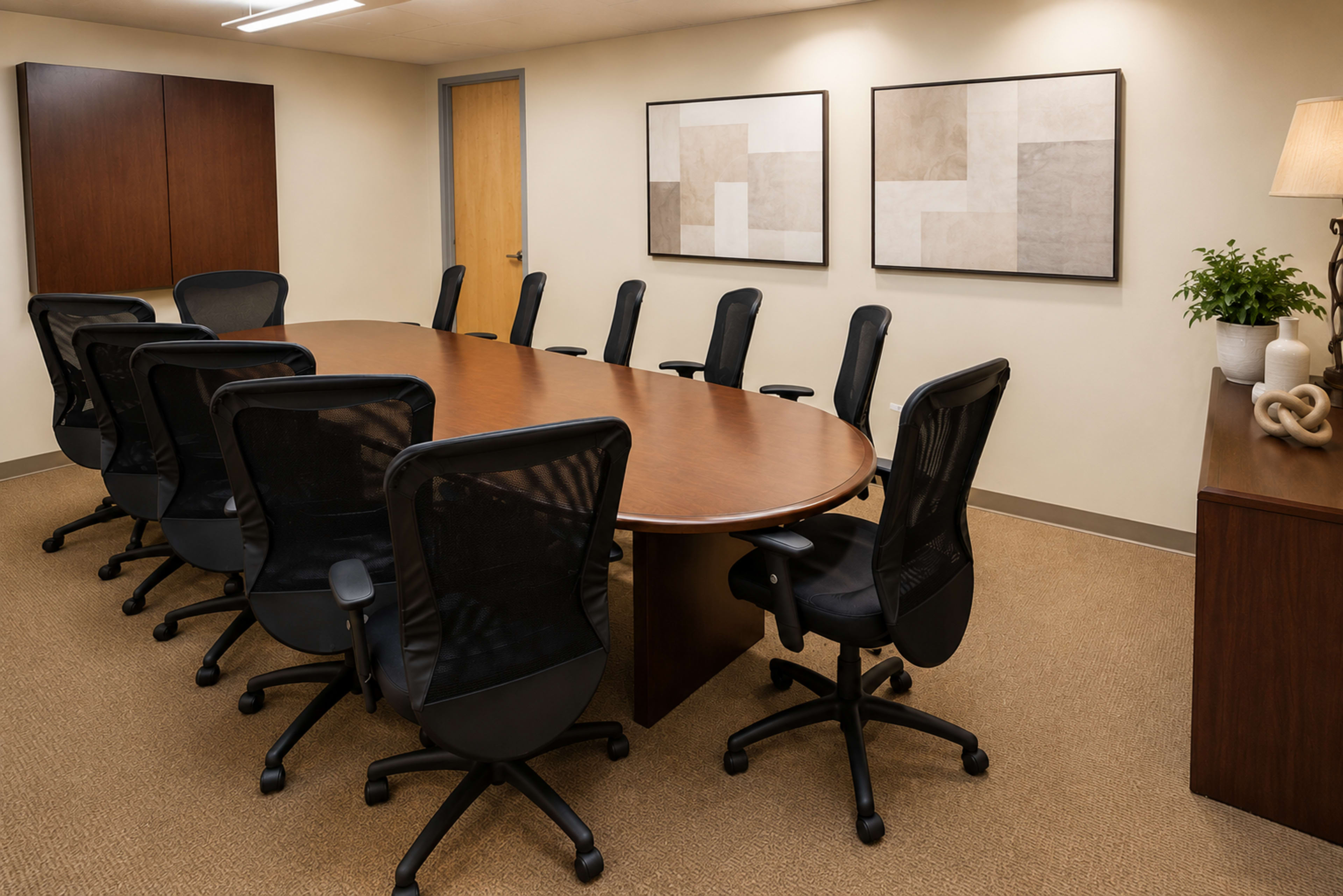 The image shows a conference room with a large oval table surrounded by several black ergonomic chairs and two framed artworks on the walls.