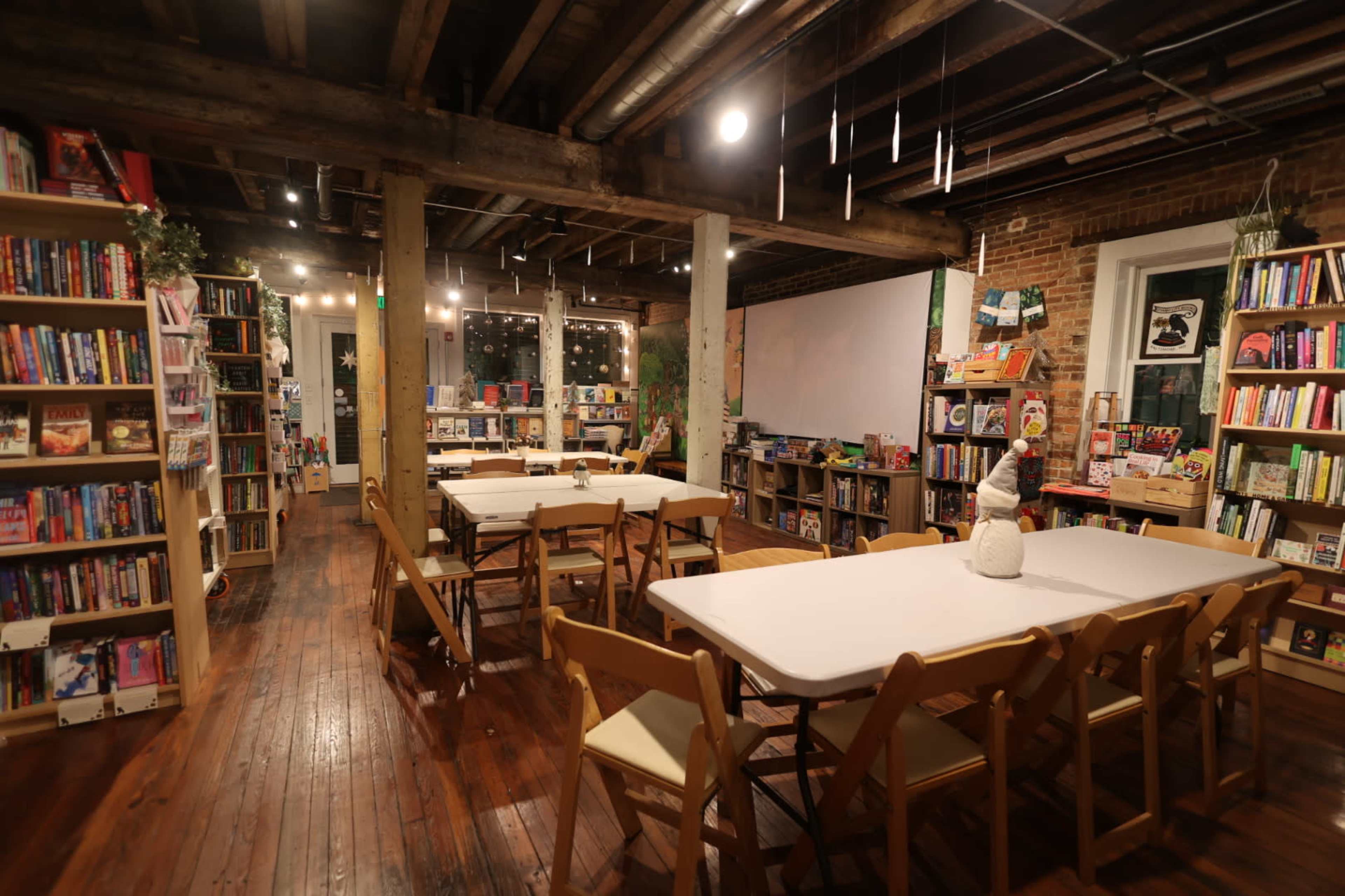 The image shows a cozy bookstore interior with wooden floors, bookshelves filled with various books, and several tables set up for reading or events.
