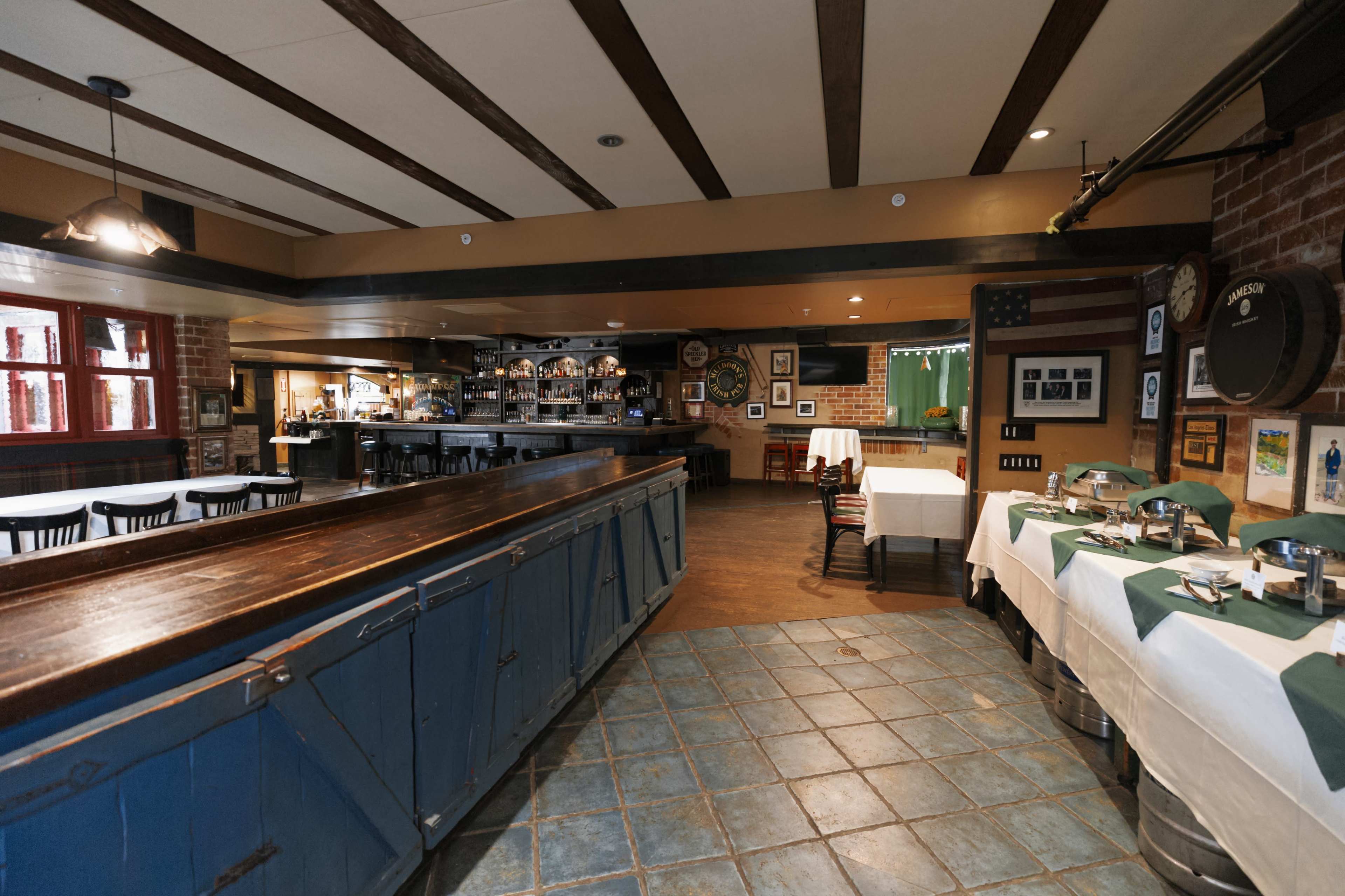 The image shows a restaurant interior featuring exposed wooden beams, a long bar counter, and tables set with green and white tablecloths.