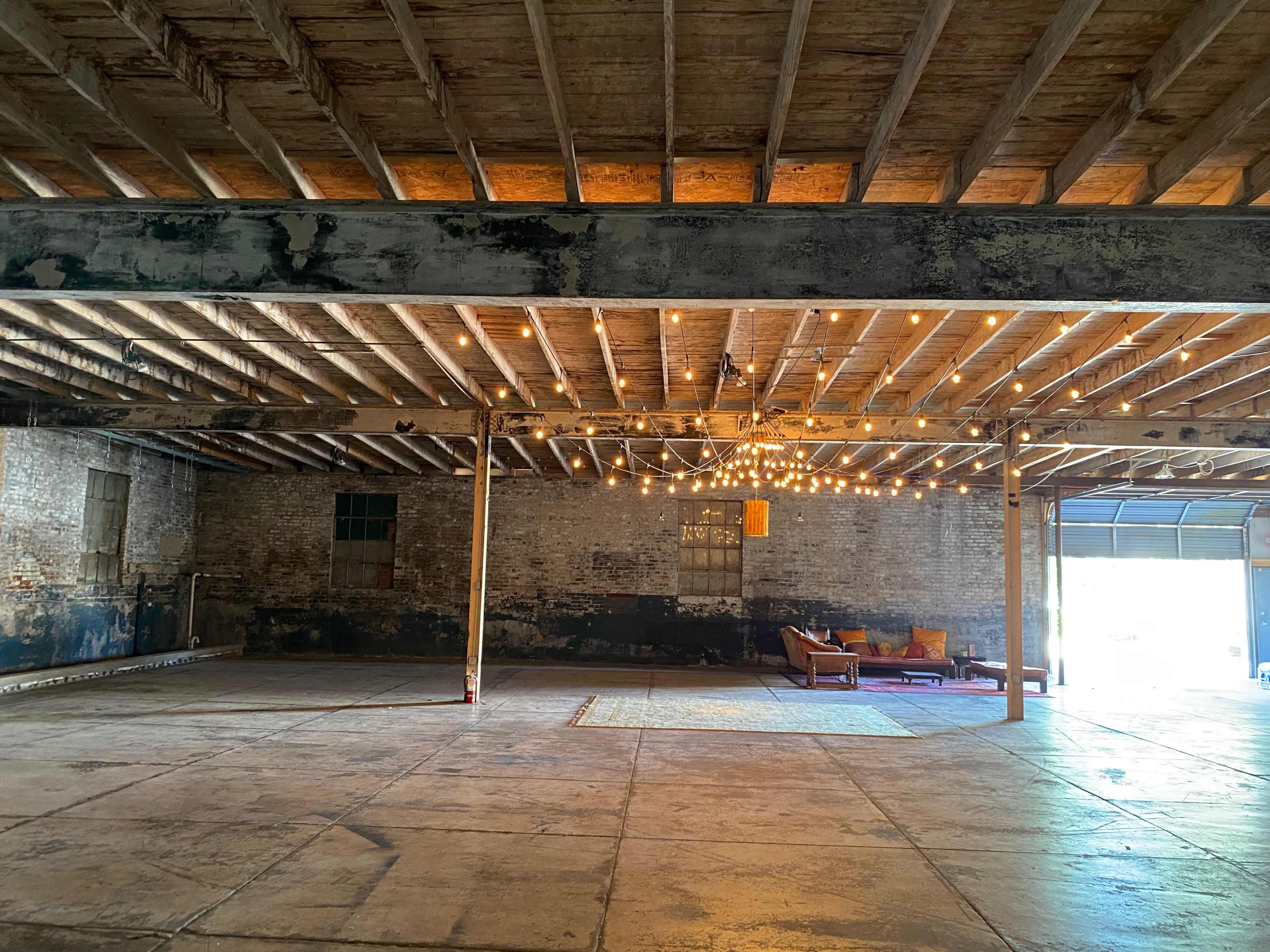A spacious industrial room with exposed wooden beams, a concrete floor, and string lights illuminating a cozy seating area.