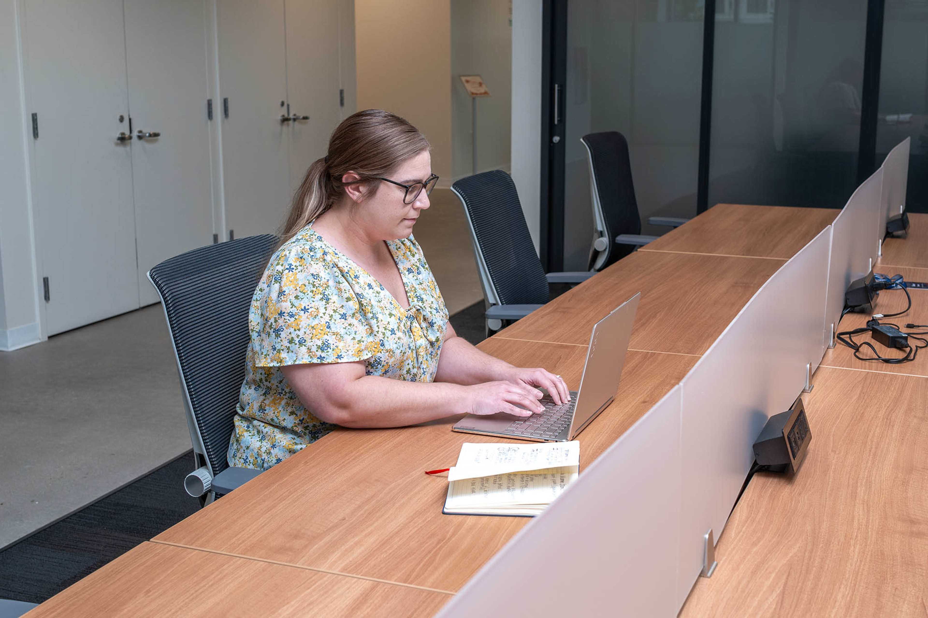 A woman sits at a large wooden table in a modern office space, typing on a laptop with a notebook and pen beside her.