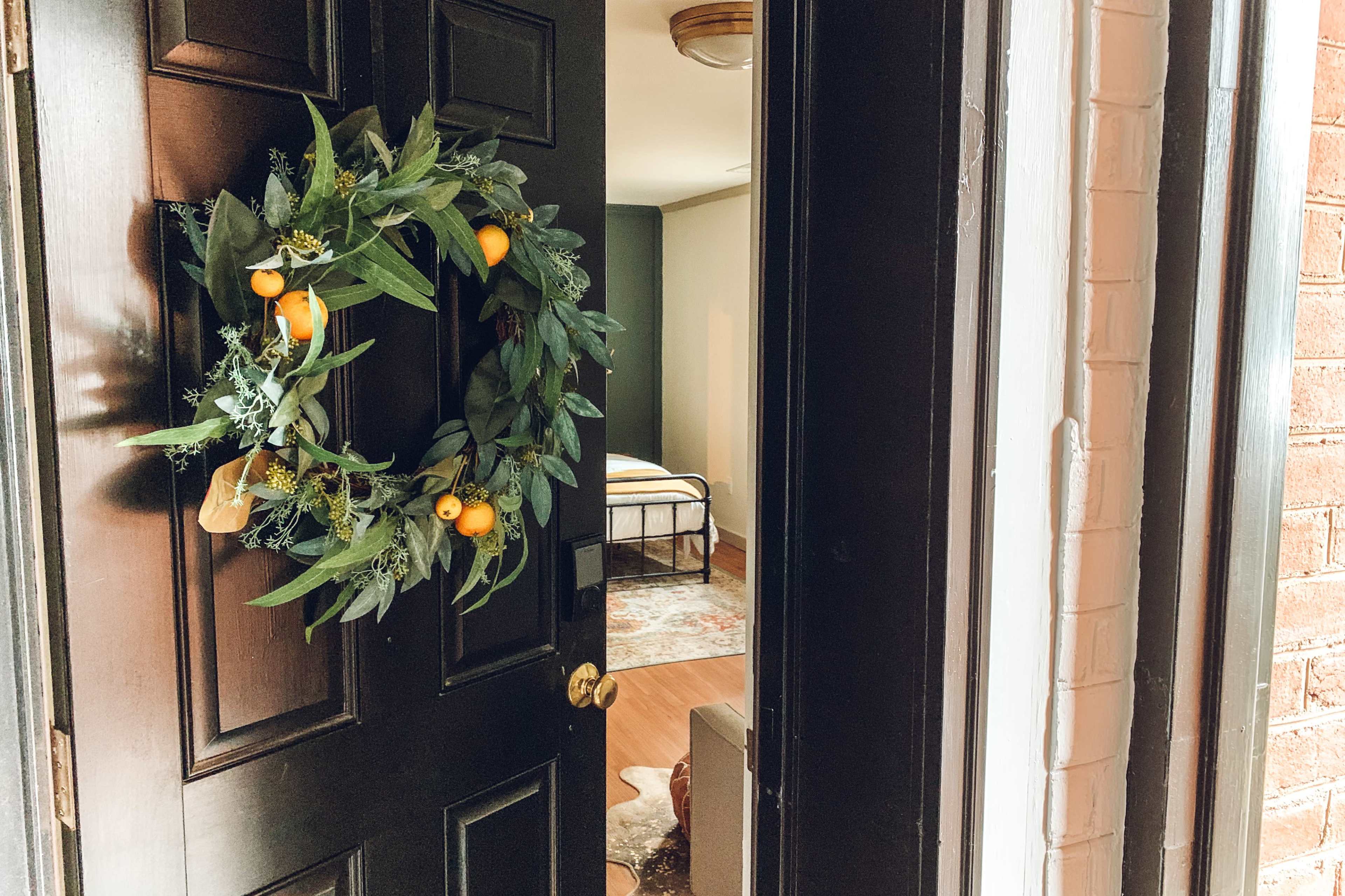 A decorative wreath with green leaves and yellow fruit hangs on a dark front door, revealing a neatly arranged interior room in the background.