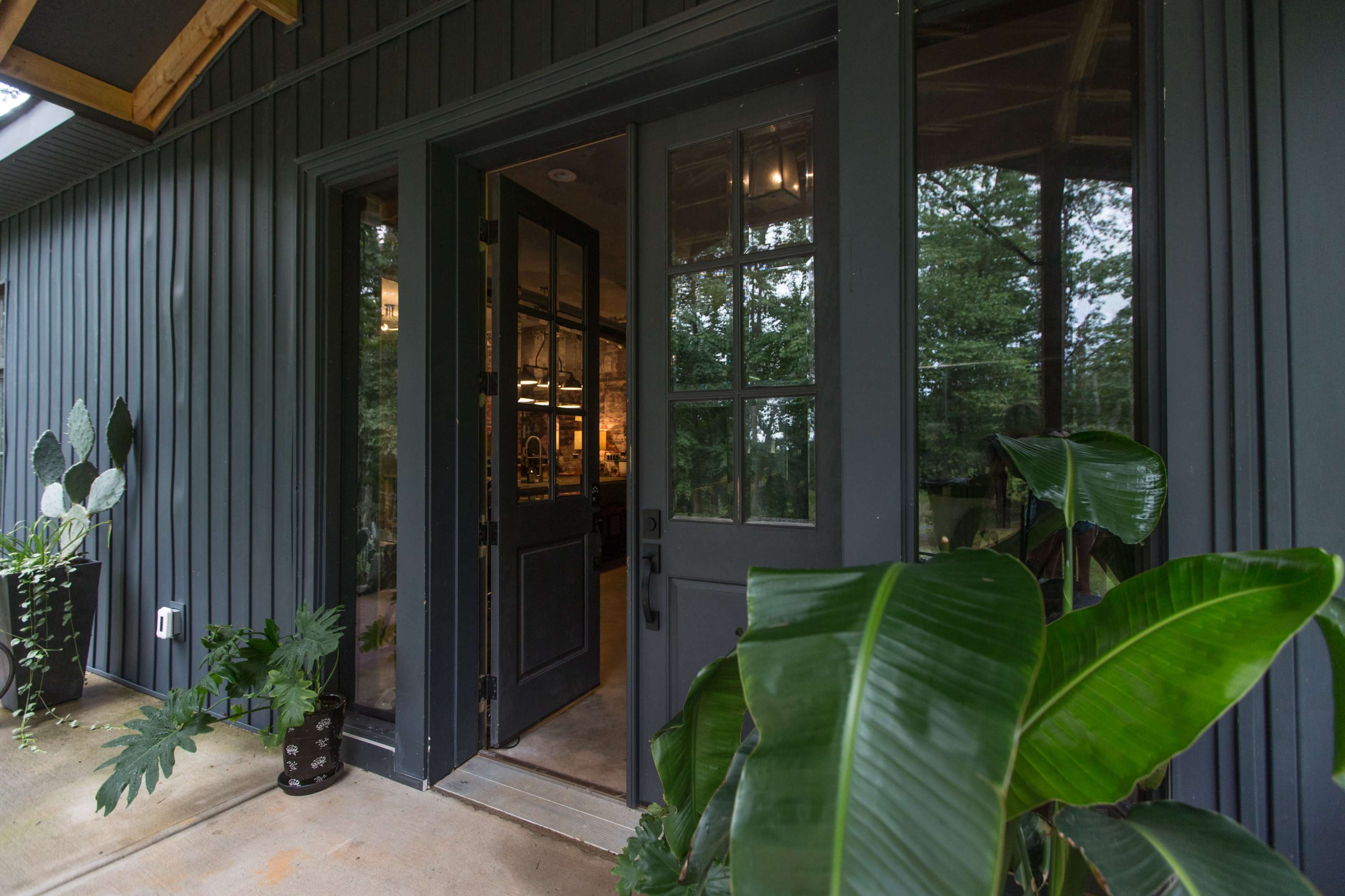 The image shows a dark-paneled entrance with a glass-paneled door, surrounded by greenery and leading into a well-lit interior.