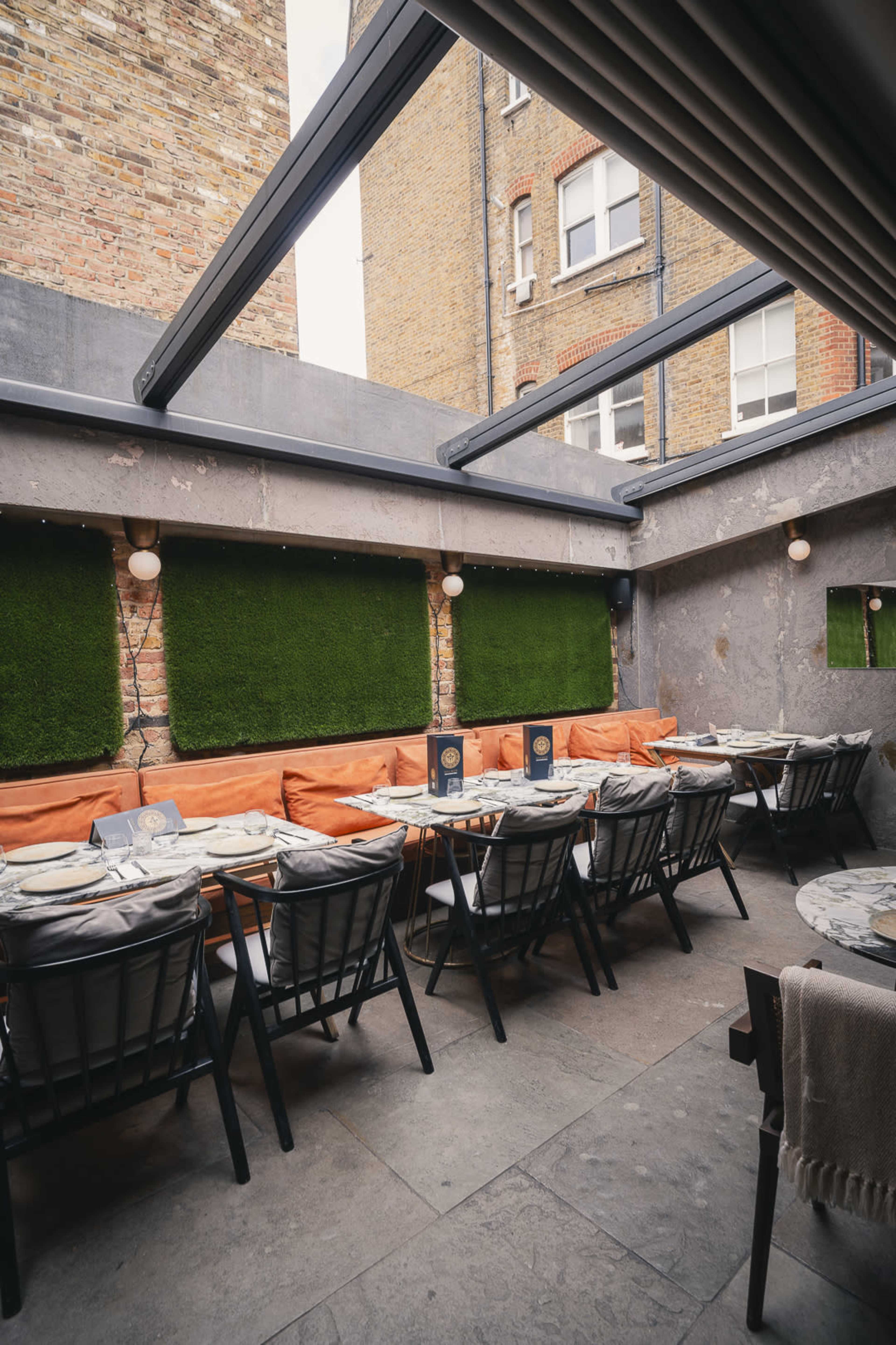 The image shows an outdoor dining area with orange cushions on the benches against green wall panels and tables arranged neatly for guests.