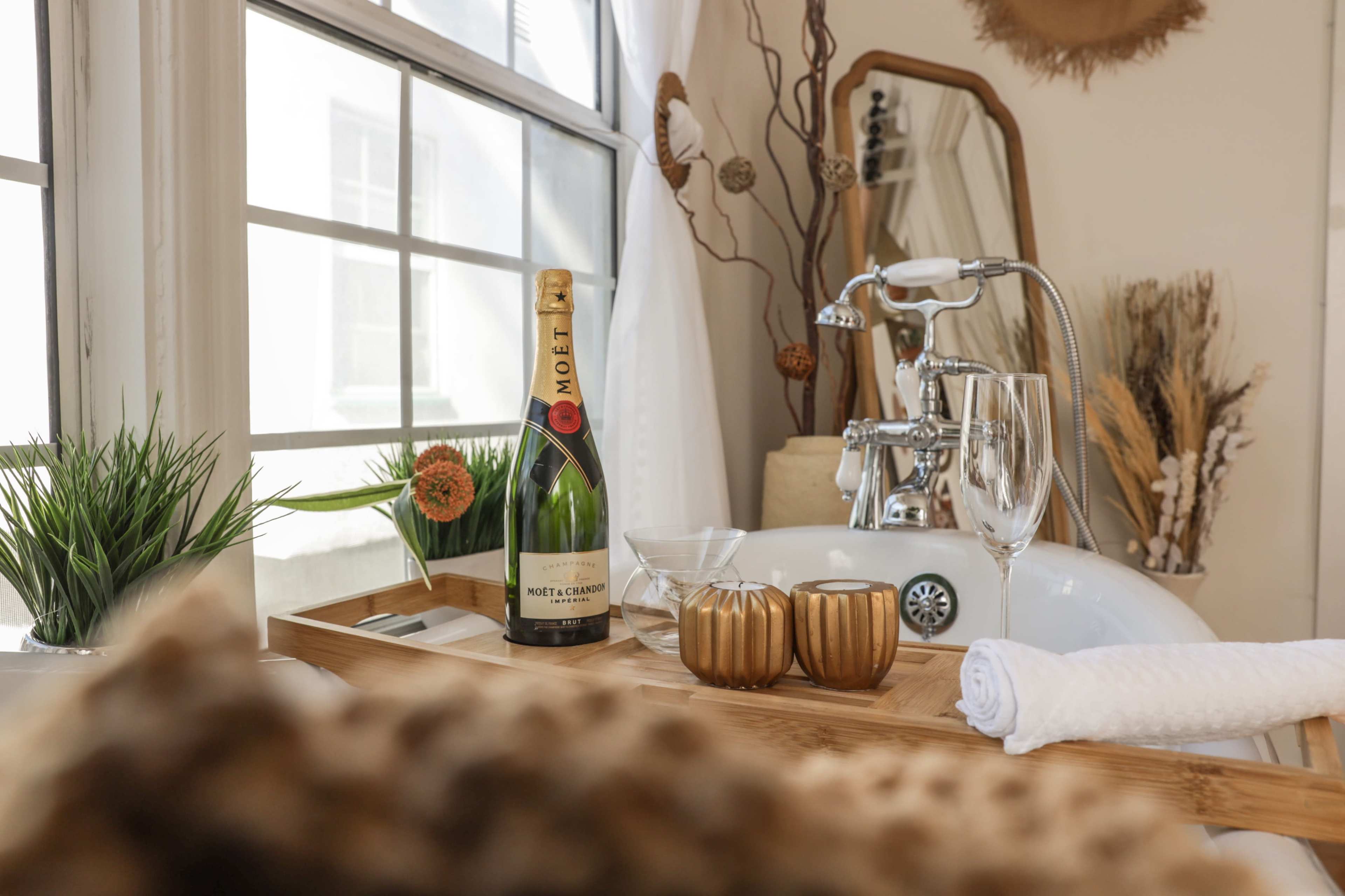A wooden tray holds a bottle of champagne, two glasses, and decorative items beside a vintage bathtub with a large mirror and window in the background.