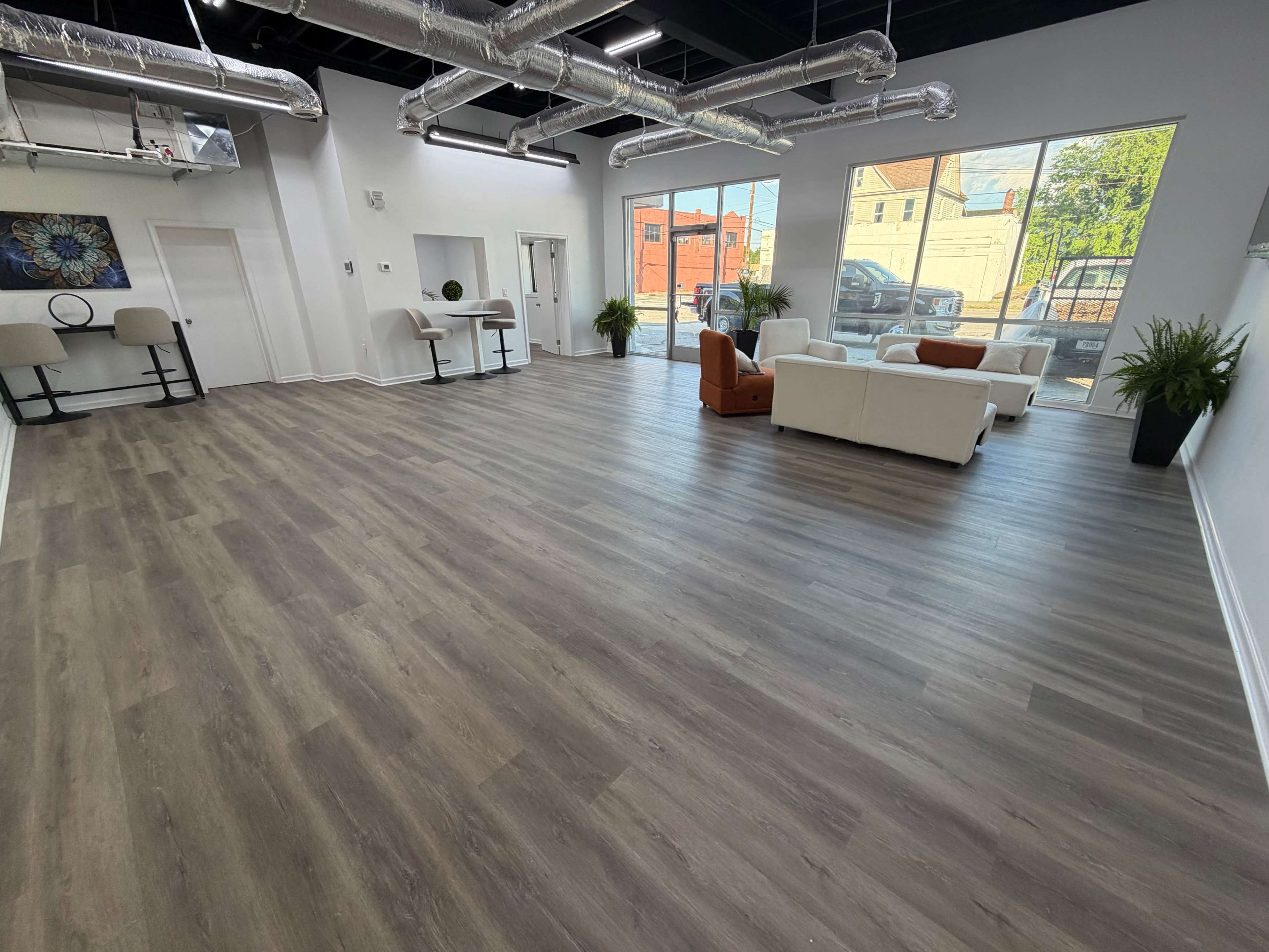 The image shows a spacious, modern interior with hardwood flooring, large windows, and minimal furnishings including a white sofa, two armchairs, and potted plants.