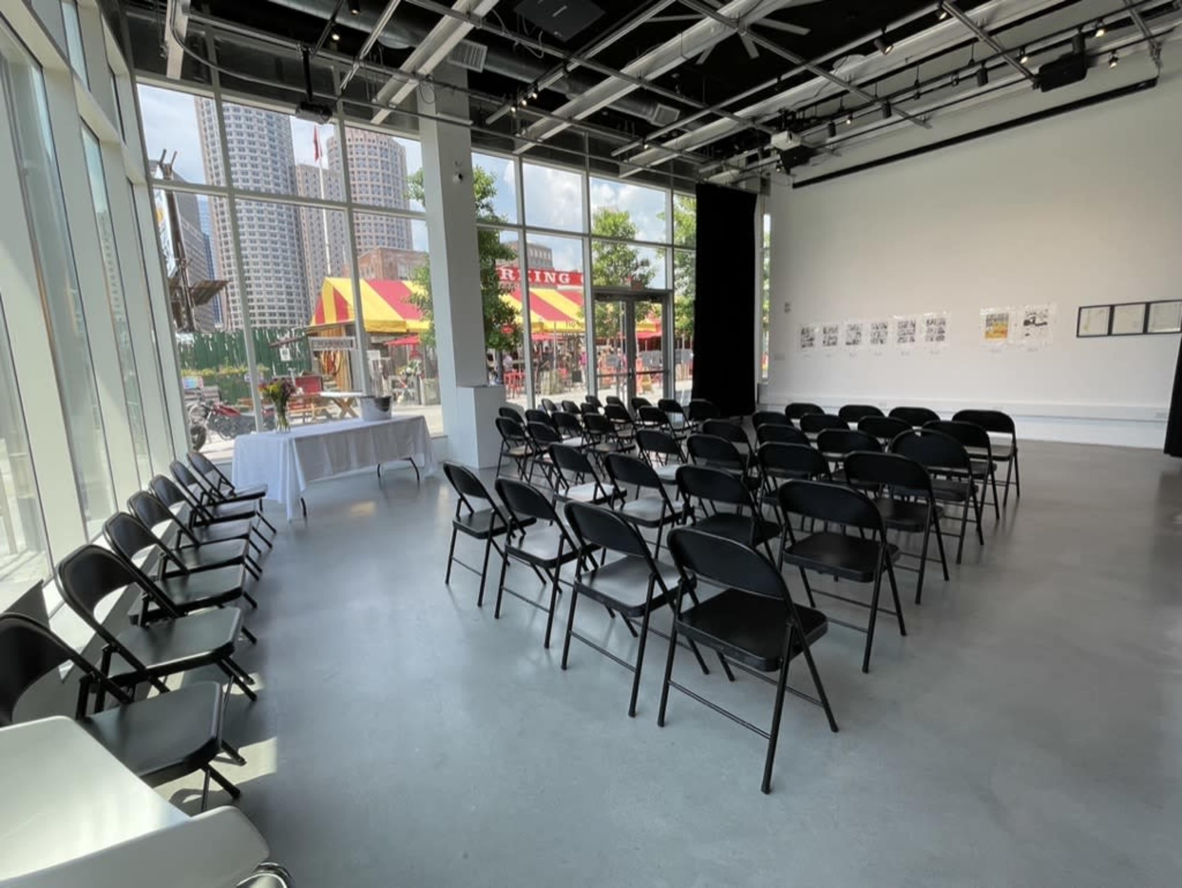 The image depicts a brightly lit gallery space with rows of black folding chairs arranged facing a blank wall, and large windows showcasing a food vendor outside.