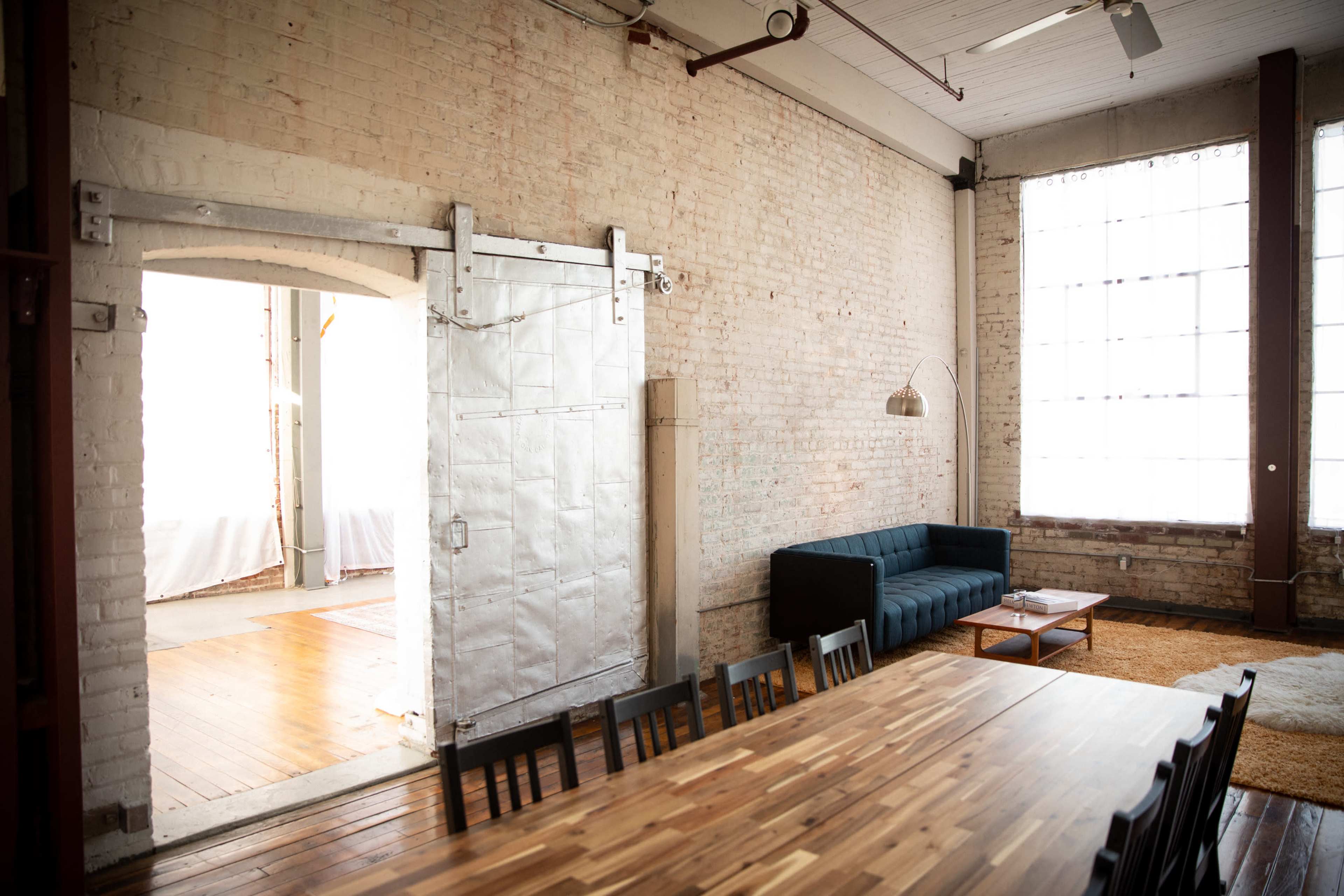 A spacious, industrial-style room with exposed brick walls, a large wooden dining table, a blue couch, and a sliding door opening to another area.
