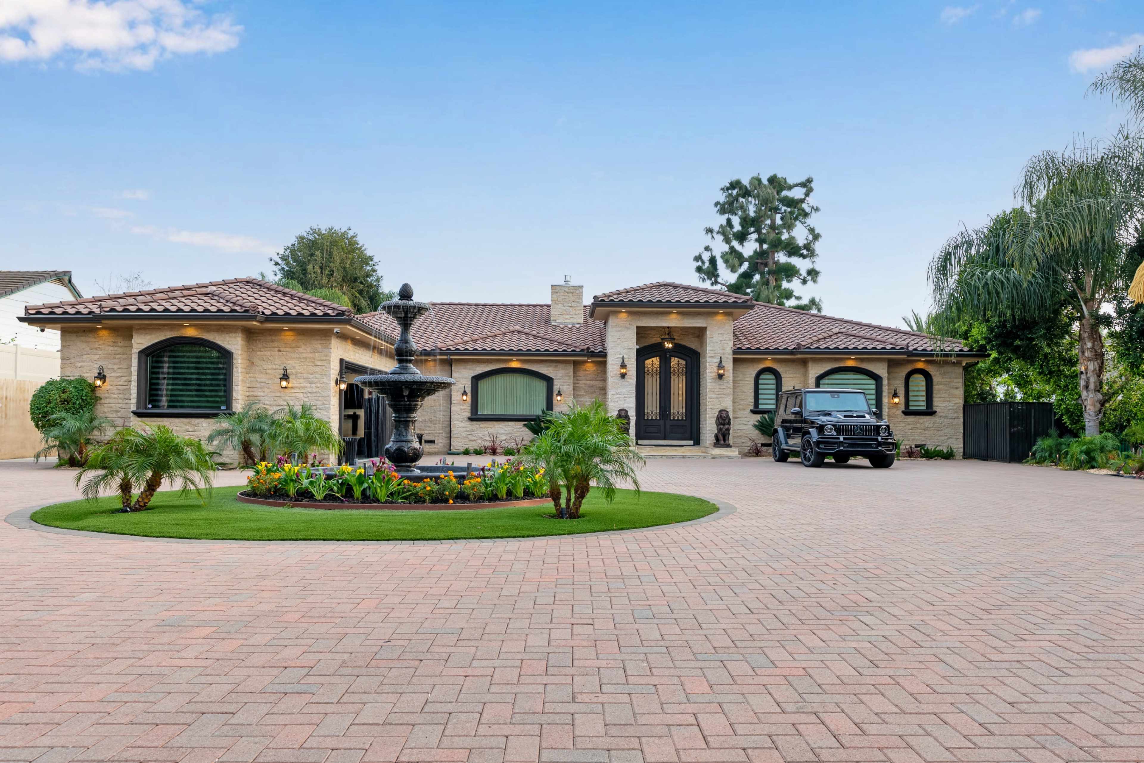 The image shows a luxury single-story home with a brick driveway, a central fountain, and decorative landscaping.