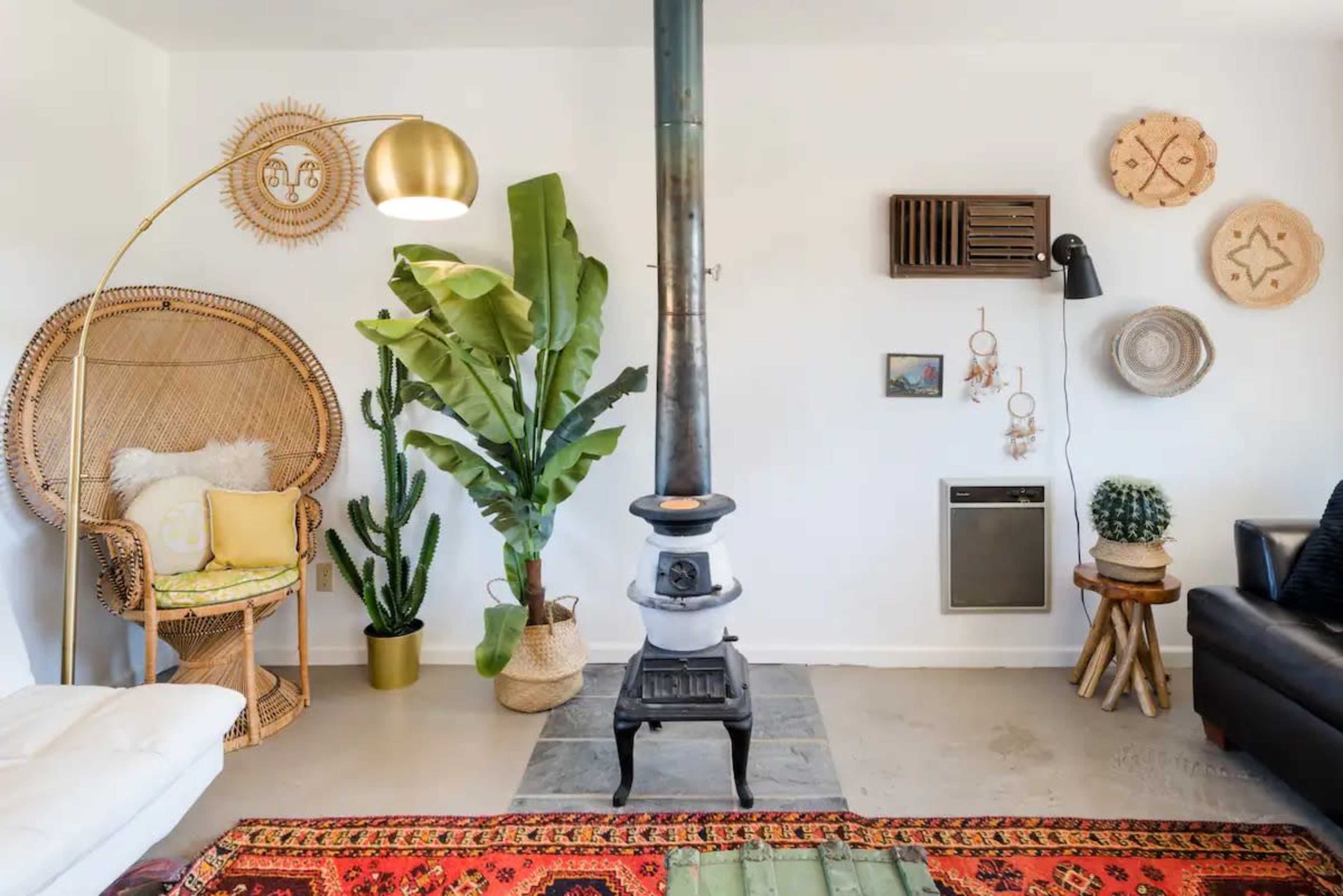 A cozy living room features a wood-burning stove, an ornate floor lamp, various plants, and decorative wall hangings.