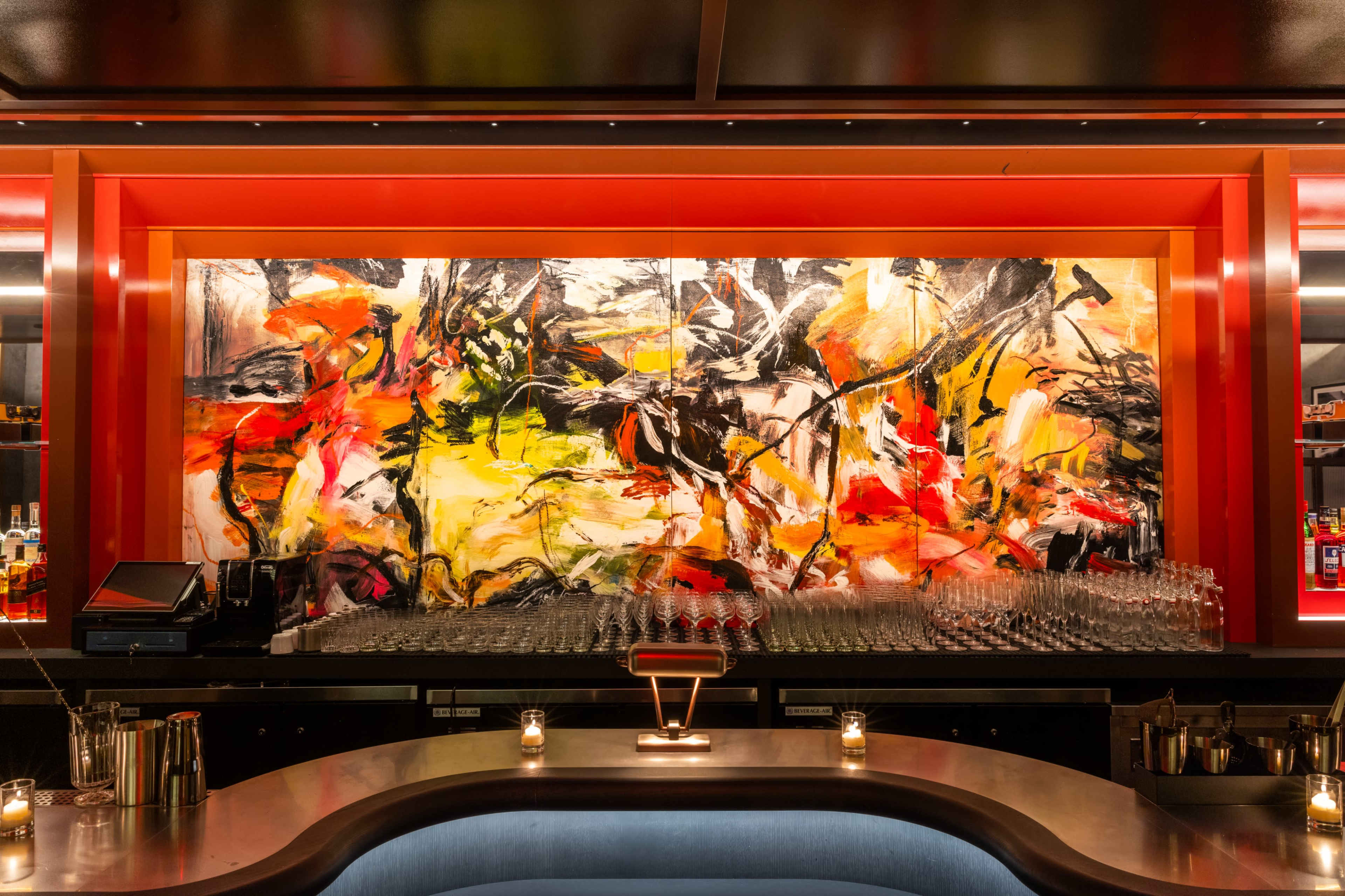 A brightly colored abstract painting hangs on the wall behind a well-stocked bar with glassware arranged along the counter.