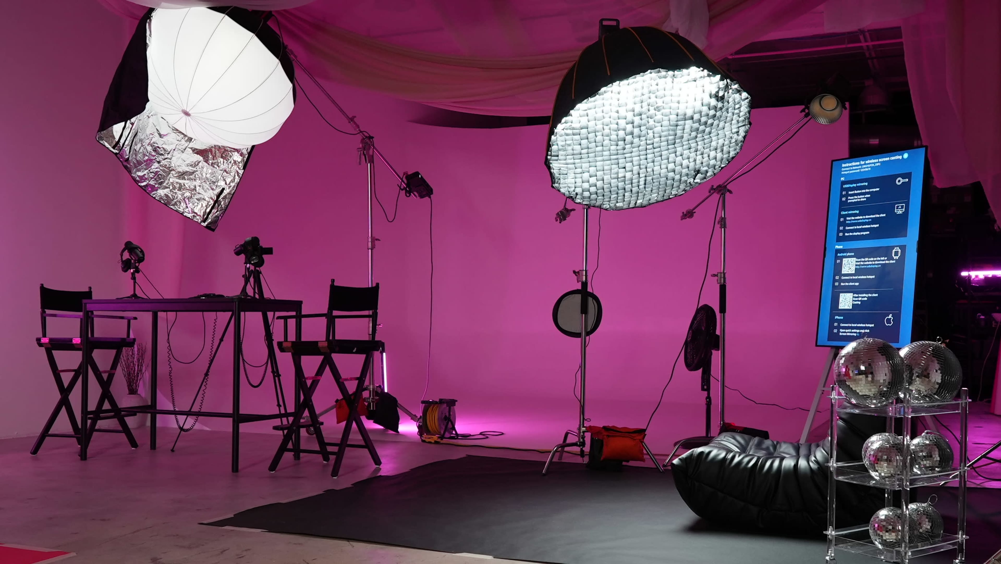 A photography studio setup with lighting equipment, black chairs, and a screen displaying information against a pink backdrop.