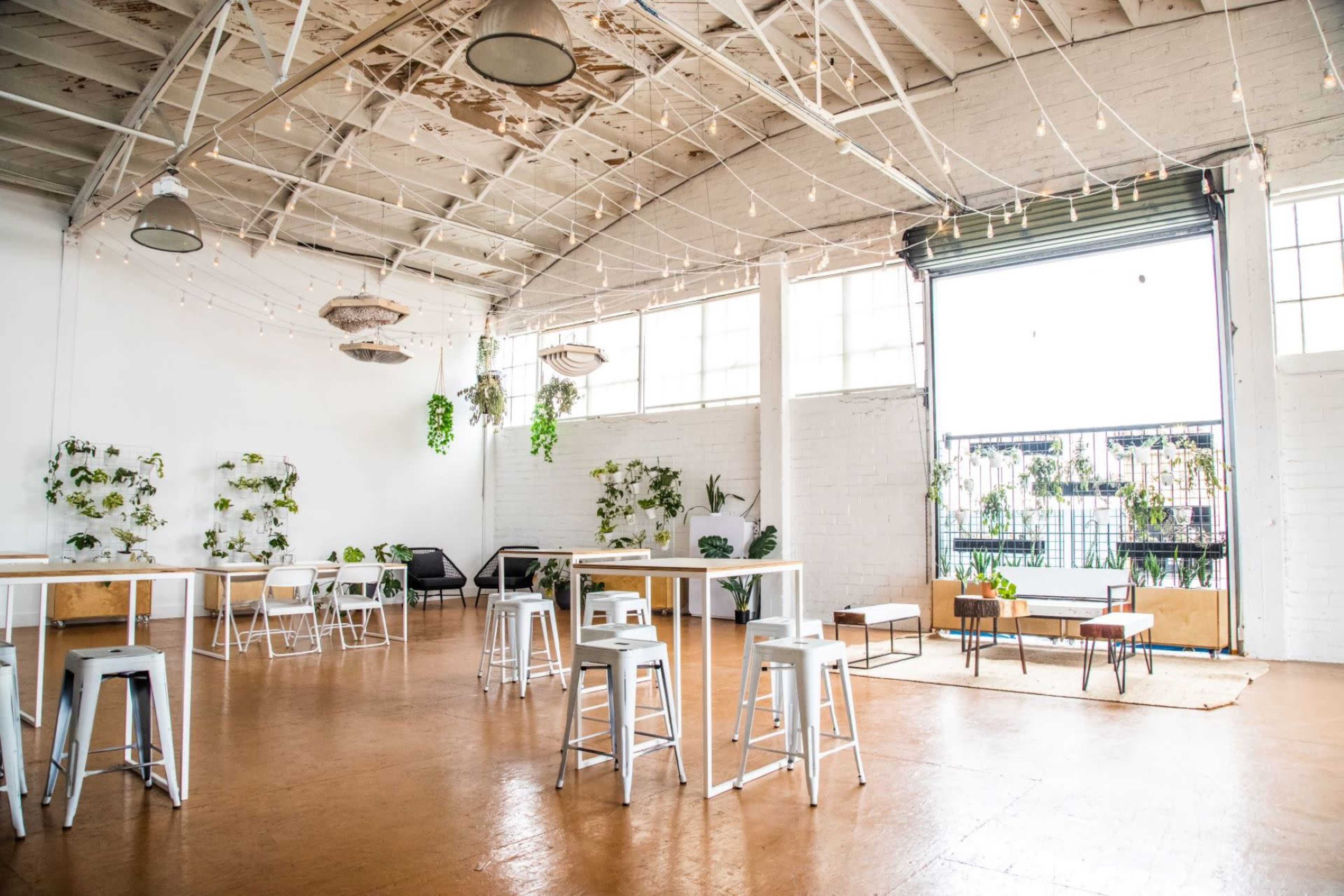 A spacious, well-lit interior with a minimalist design featuring white walls, metal tables, and seating, along with plants and large windows.