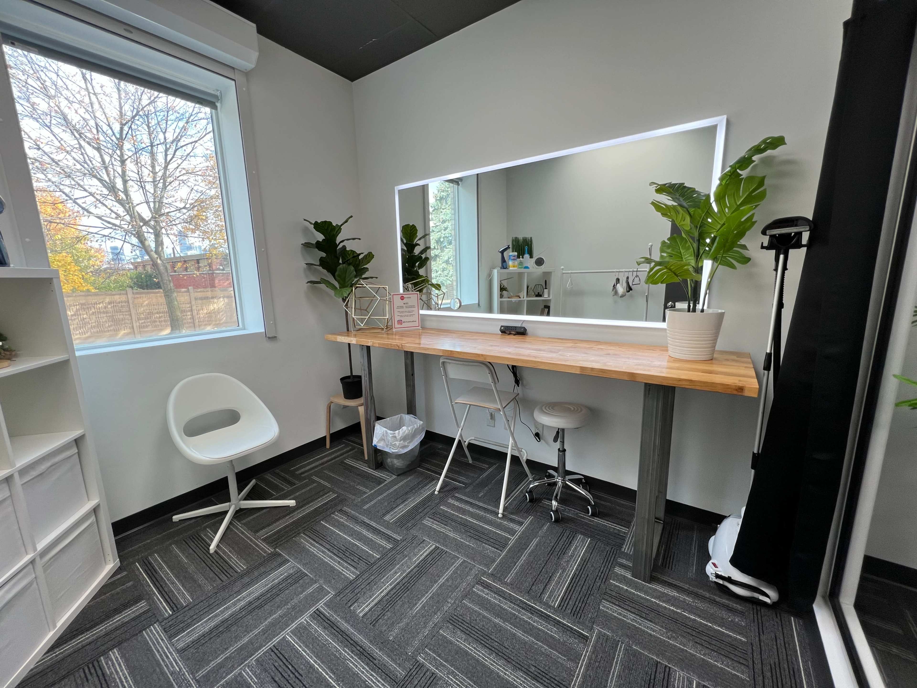 A bright, minimalist space featuring a sleek wooden table with a mirror, two chairs, and potted plants against light-colored walls.