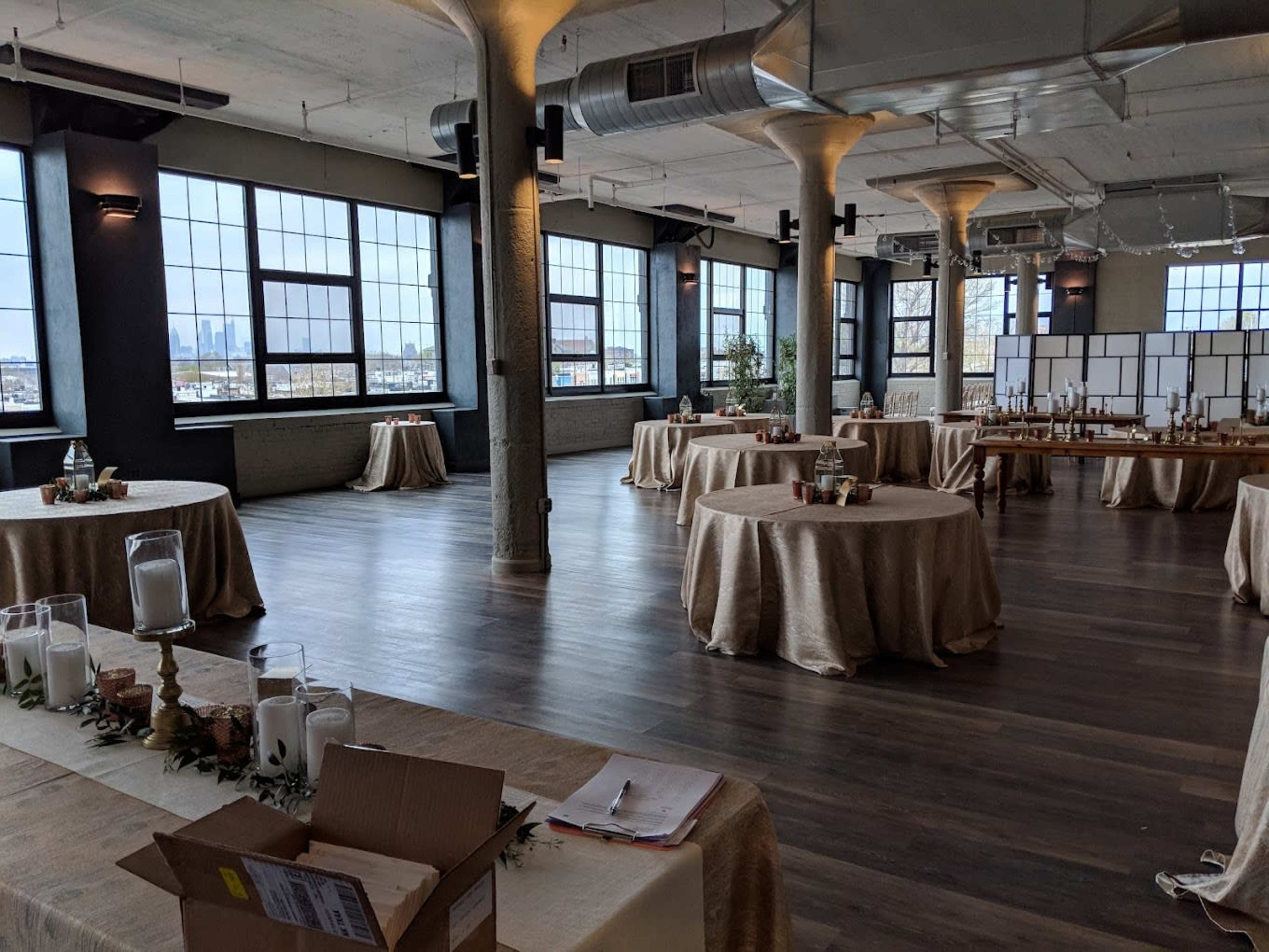 The scene shows a spacious, partially decorated event venue with several round tables covered in beige tablecloths and large windows offering a view of the city skyline.