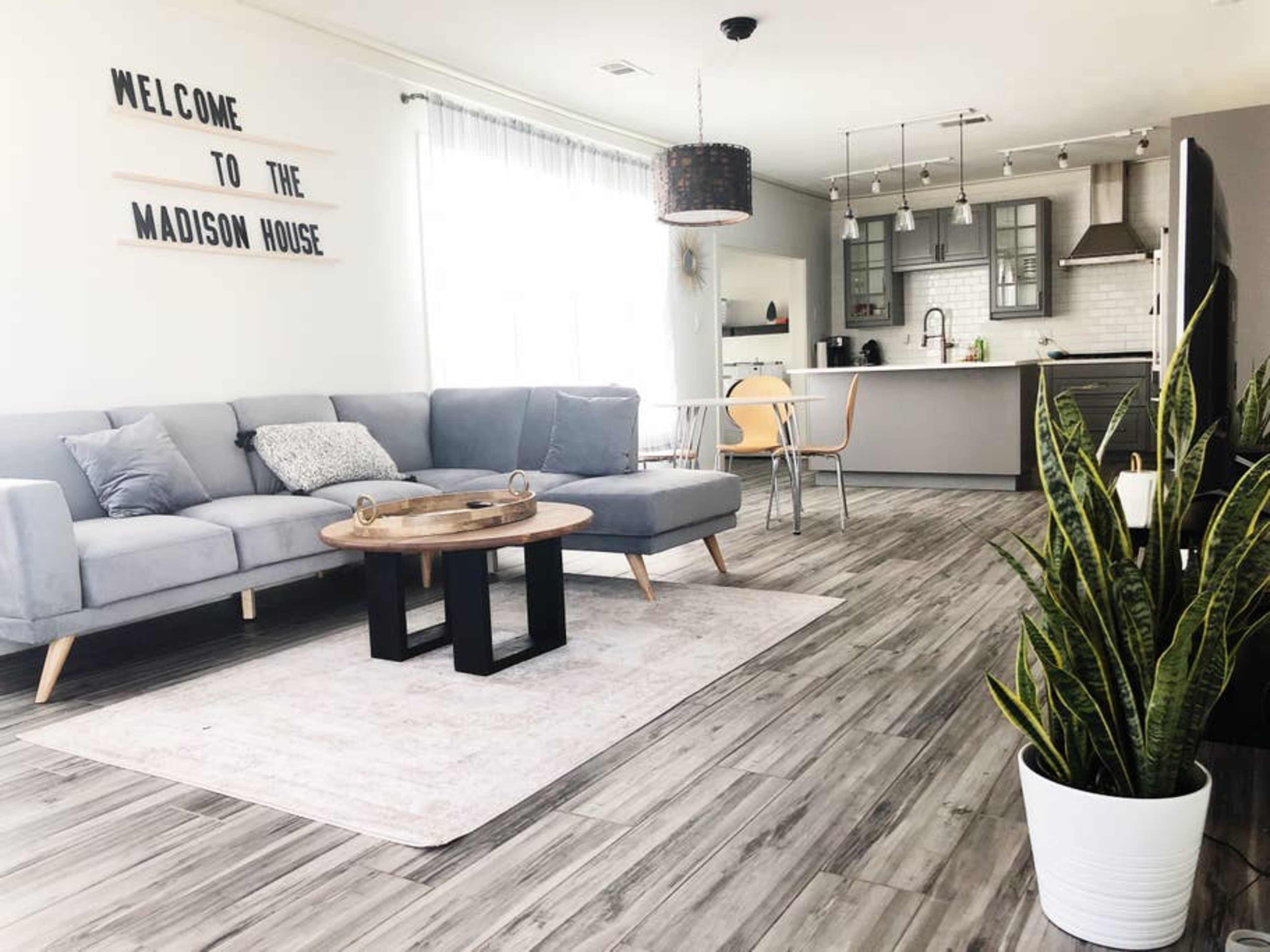 A modern living room featuring a gray sectional sofa, a round wooden coffee table, and a kitchen area in the background with stainless steel appliances.