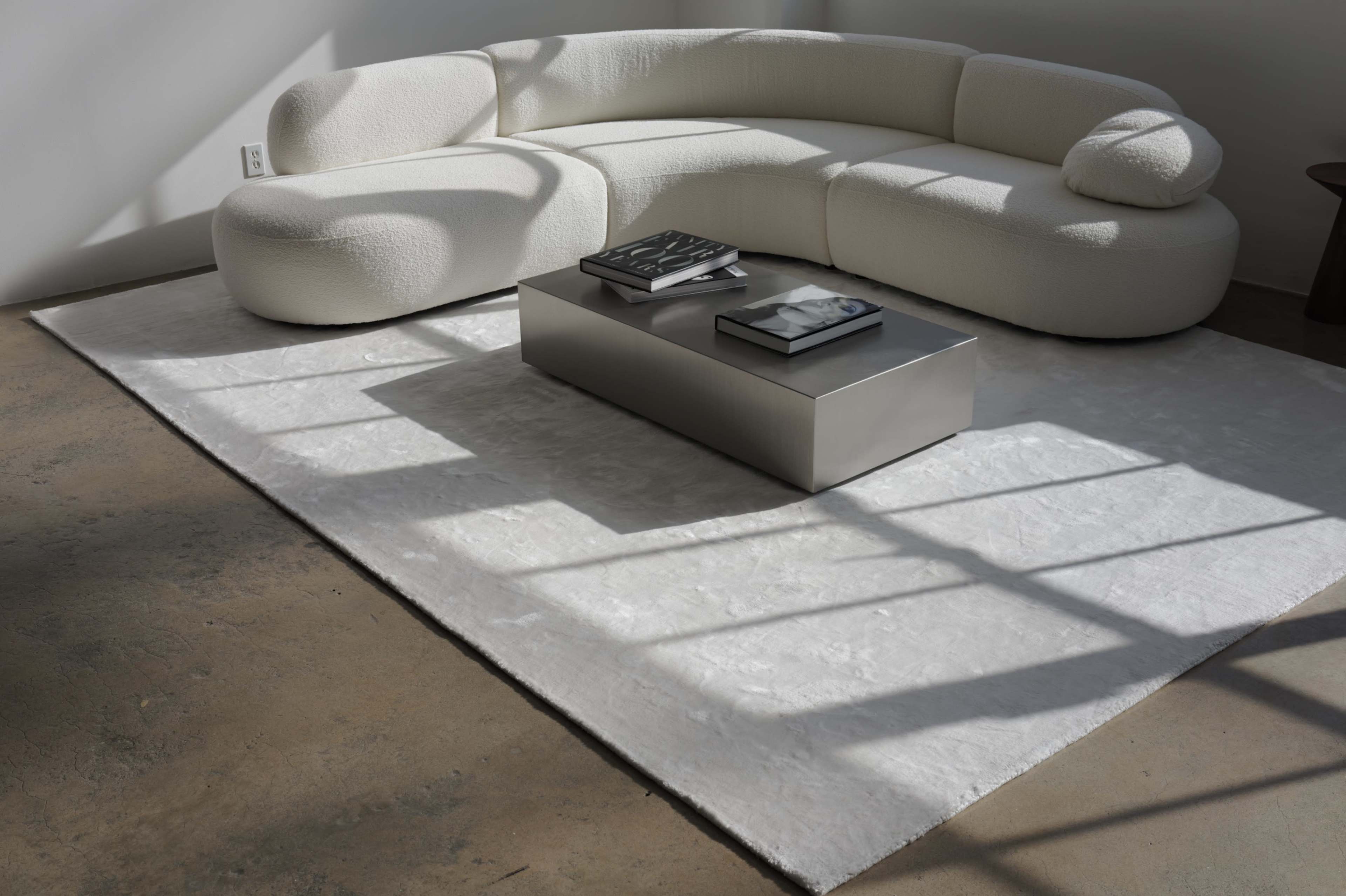 A modern, curved white sofa is positioned on a light rug next to a rectangular silver coffee table, with sunlight casting shadows on the floor.