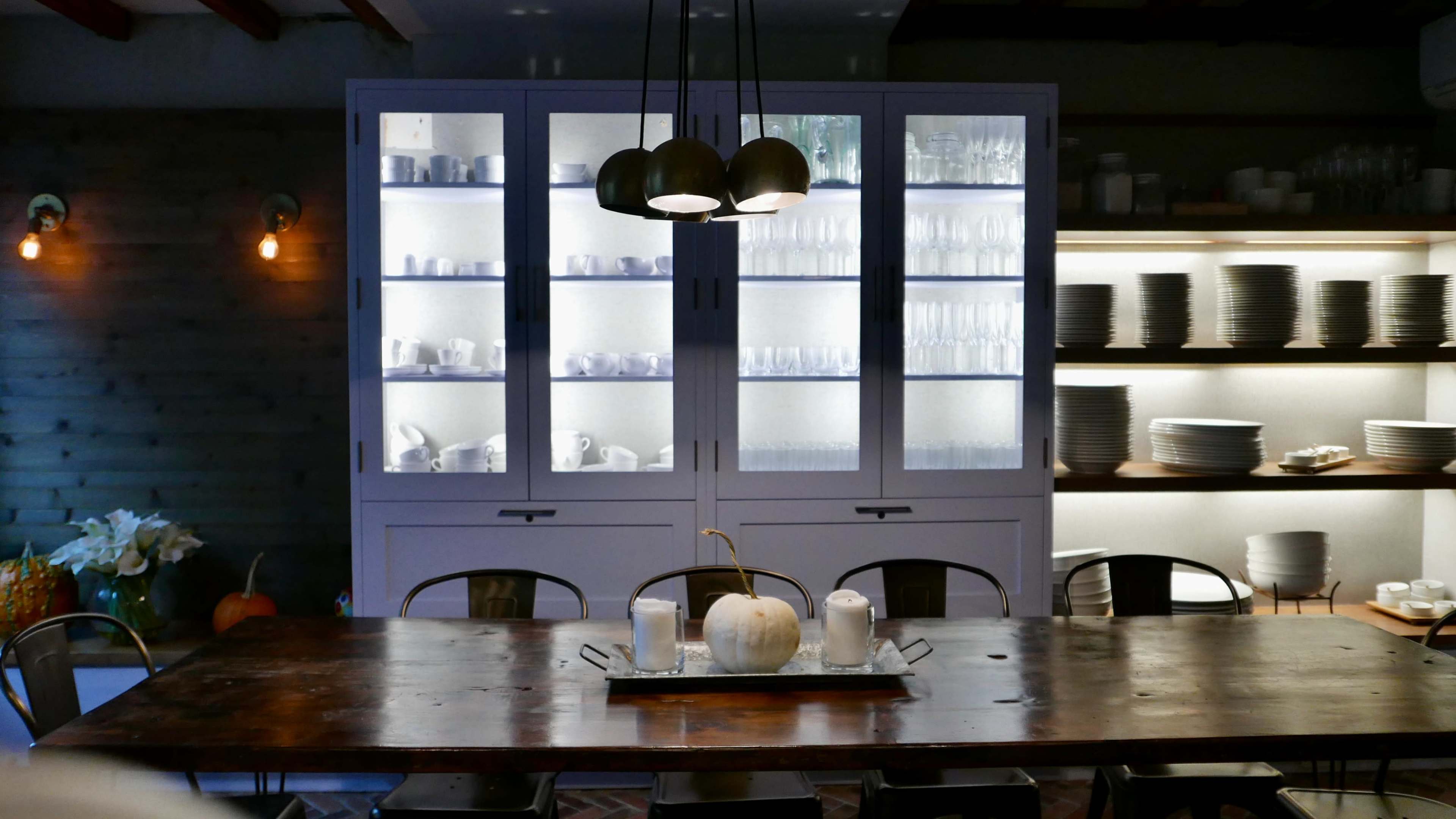 A large wooden dining table sits in front of a display cabinet filled with glassware and tableware, illuminated by pendant lights.