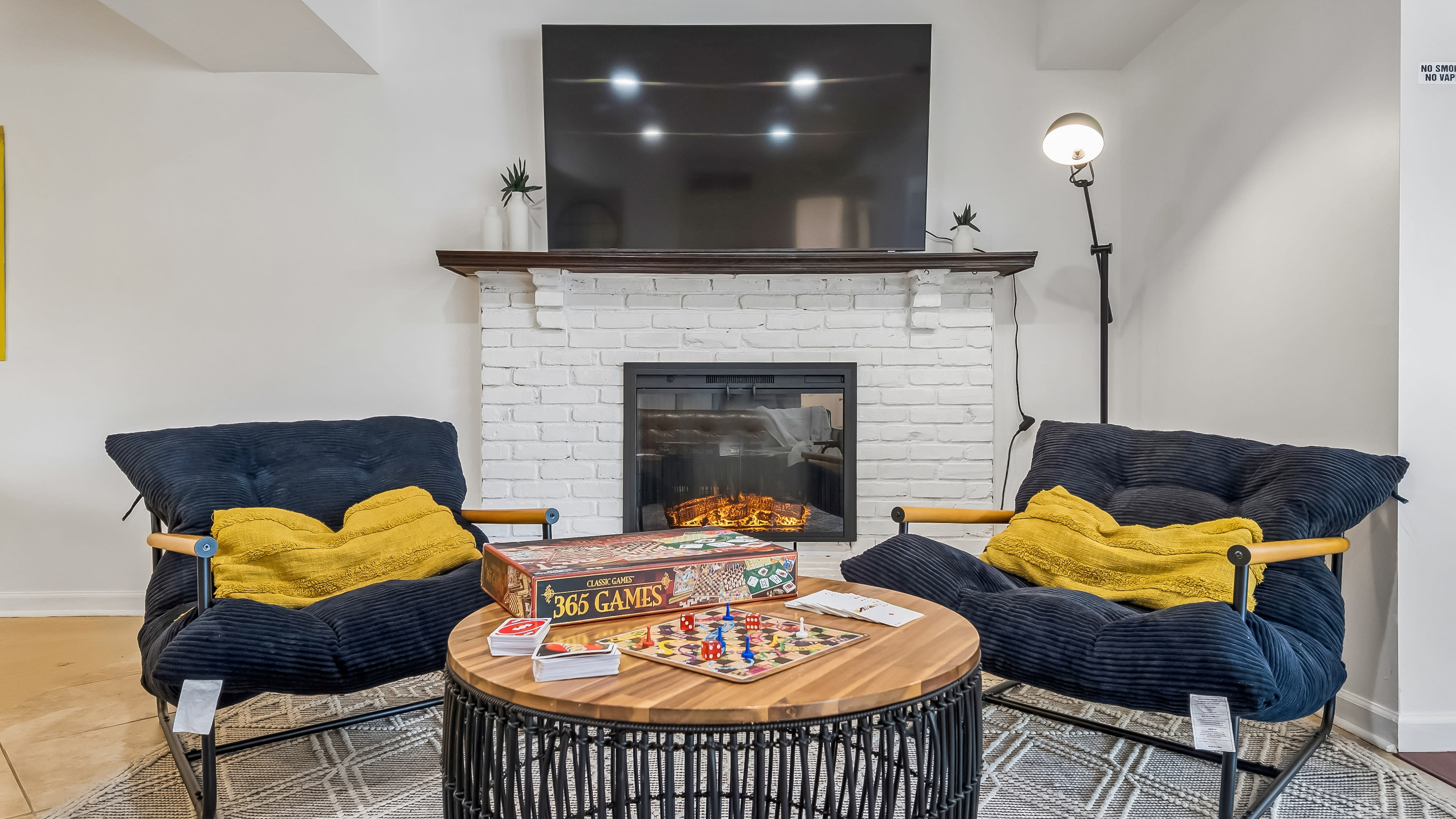 The image shows a cozy living room with two plush chairs, a round table with a board game, and a fireplace beneath a wall-mounted television.