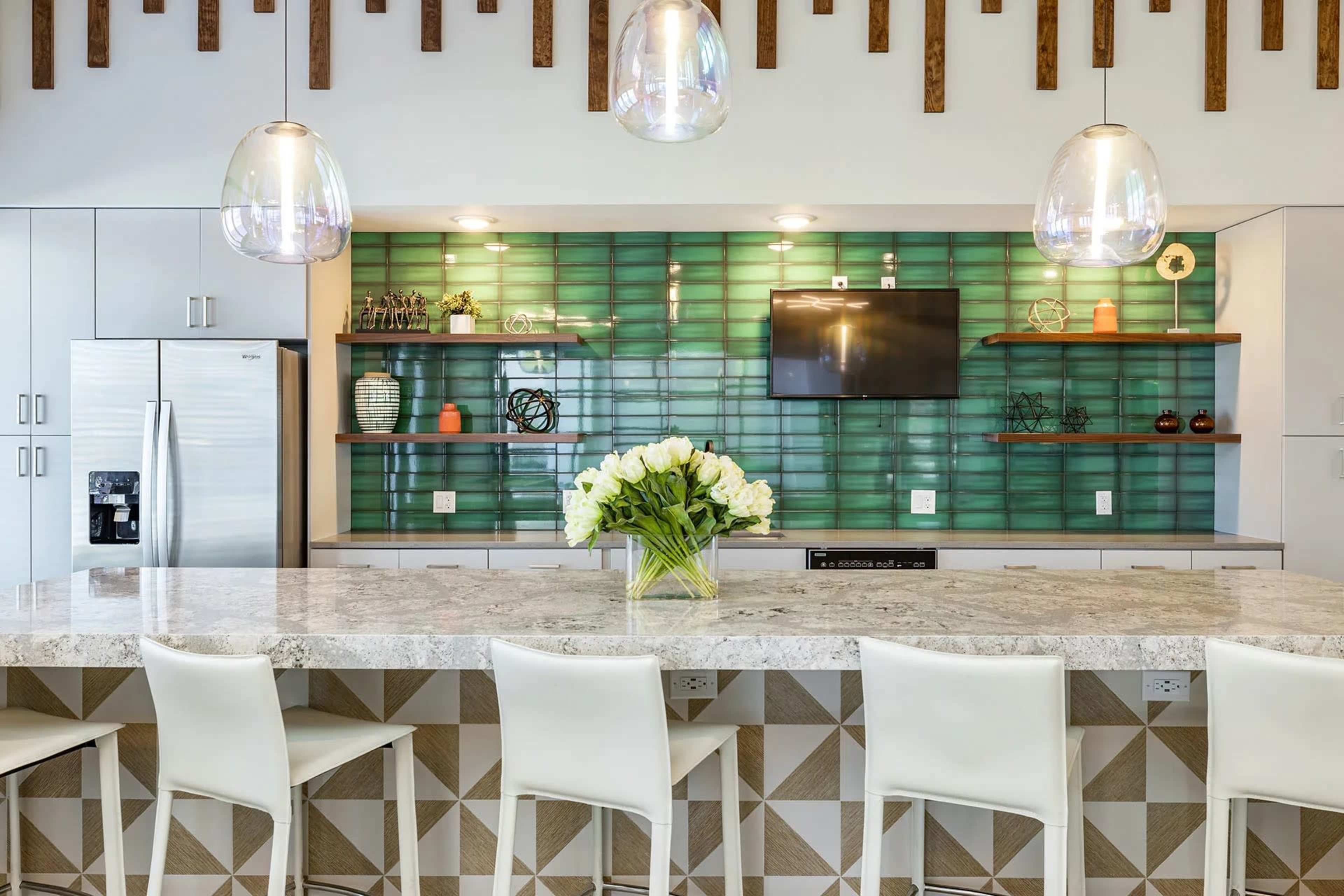 A modern kitchen features a large marble island with white barstools, a green tiled backsplash, and pendant lighting above.