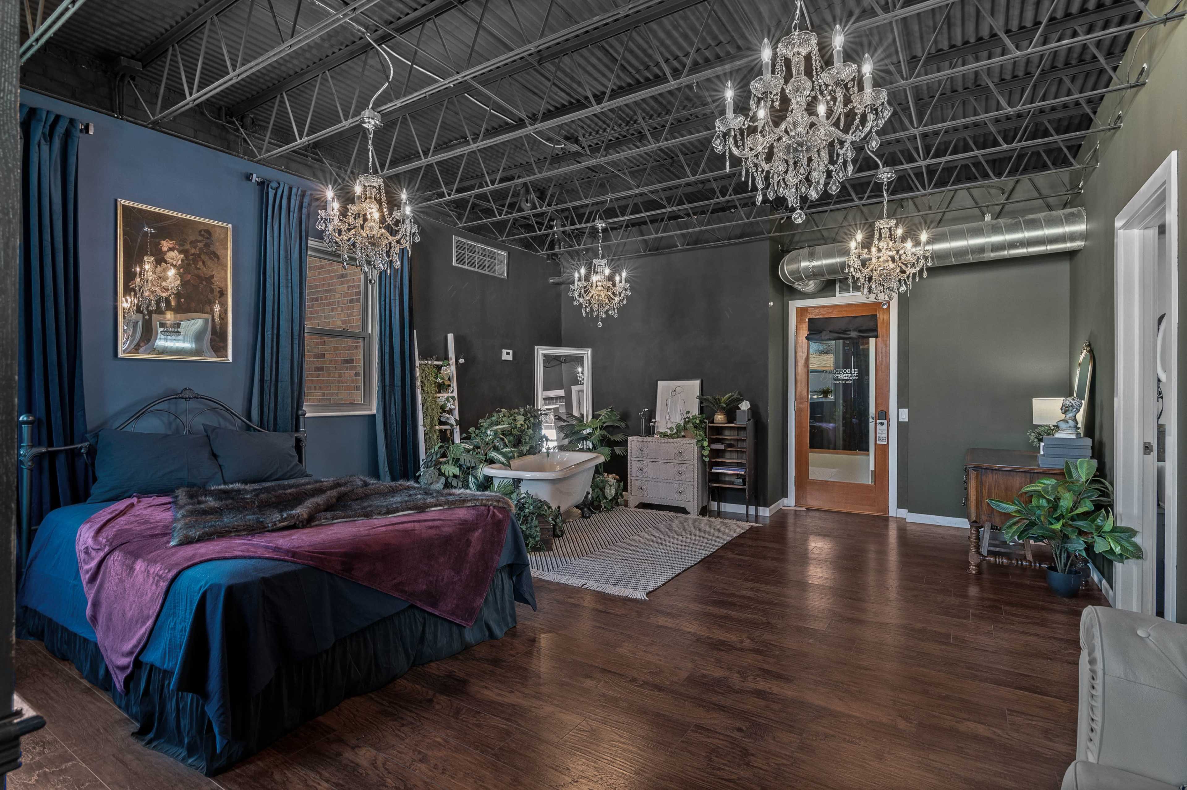 A spacious bedroom with a dark color scheme, featuring a bed, several plants, and two chandeliers hanging from the ceiling.