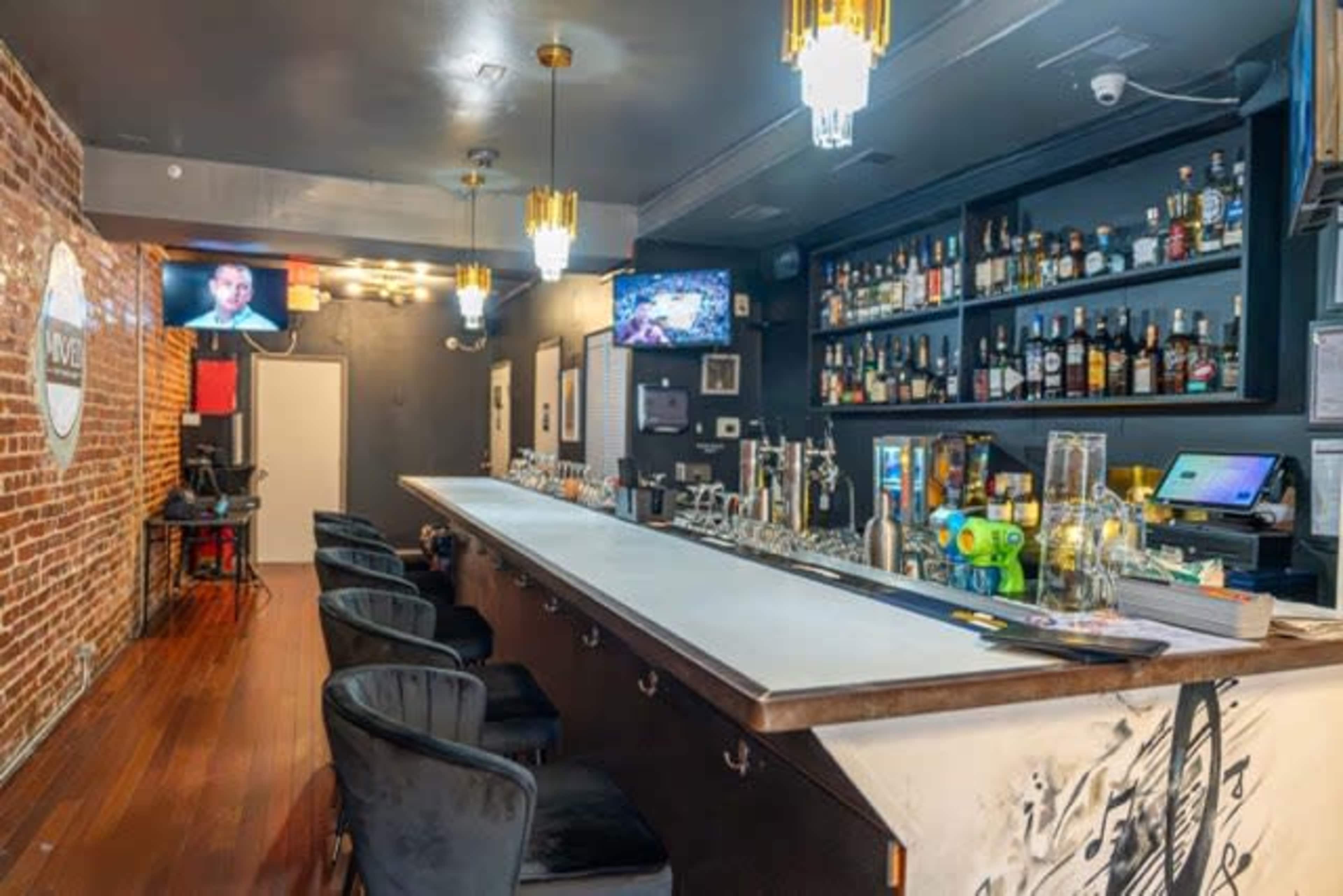 A modern bar with a wide counter, shelves stocked with various bottles, and a few bar stools along the front.