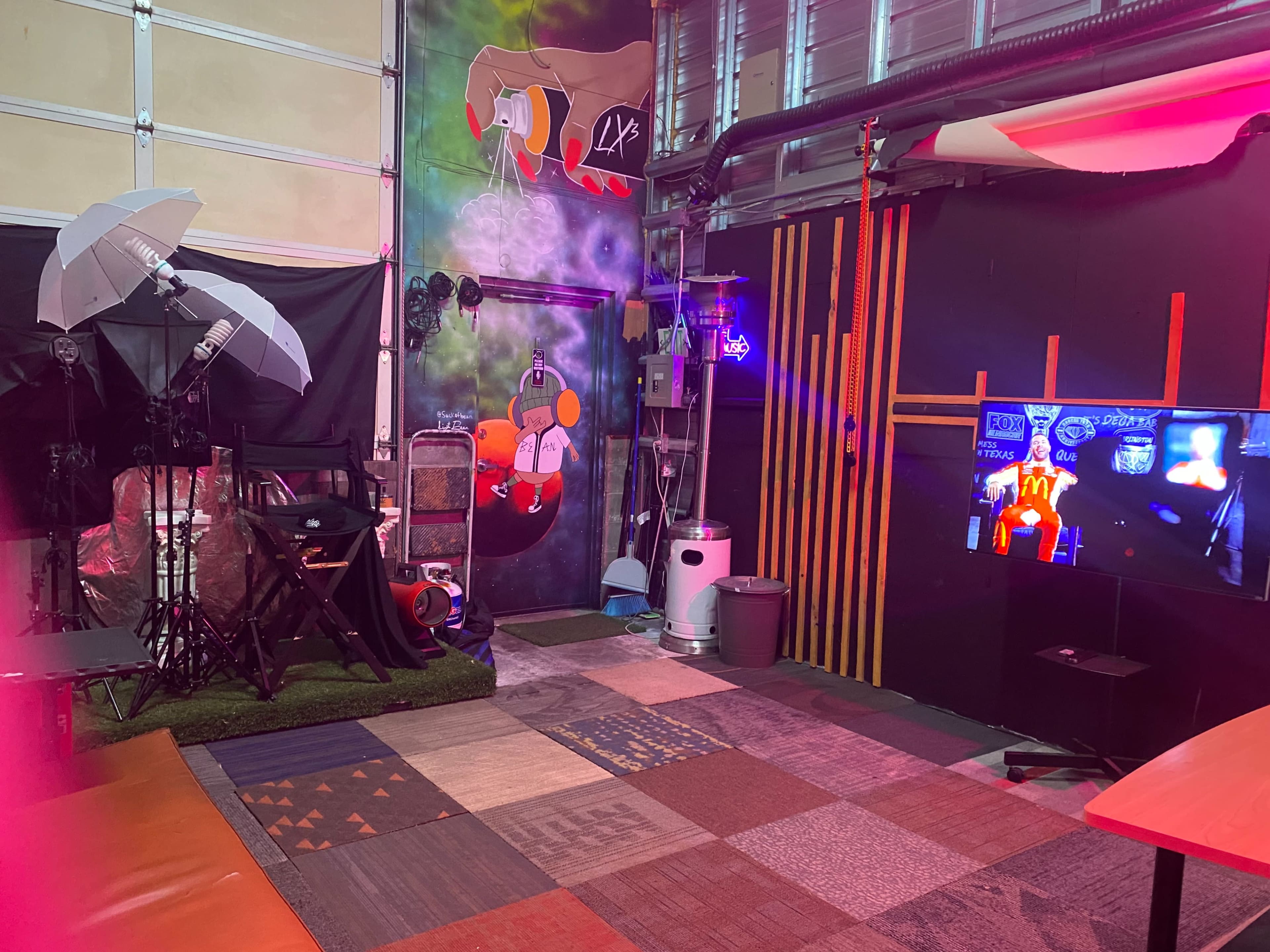 The image shows a creatively designed studio space featuring lighting equipment, a television screen, and colorful floor coverings.