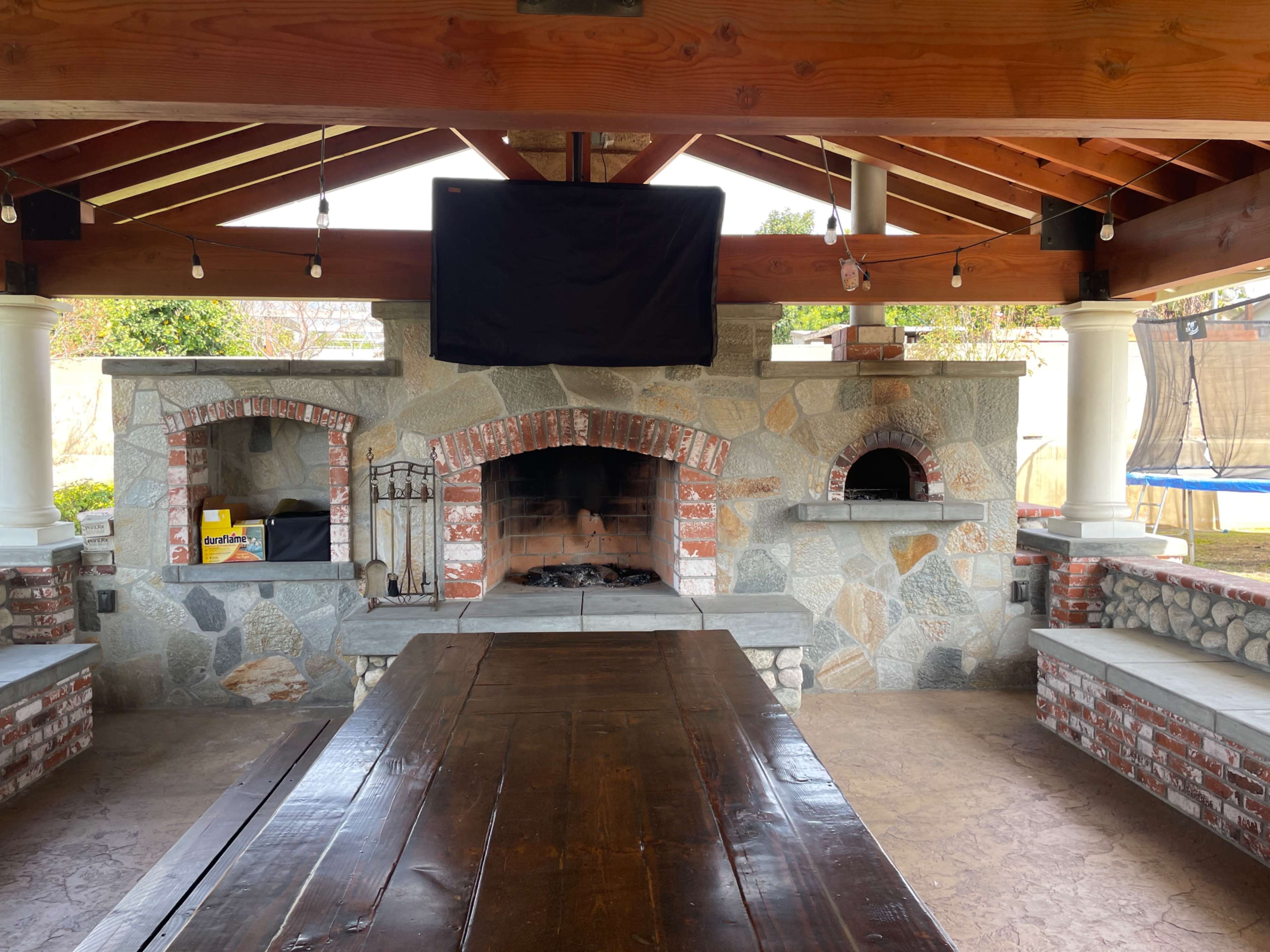 A large outdoor pavilion featuring a stone and brick fireplace with two ovens and a wooden dining table underneath a wooden ceiling.