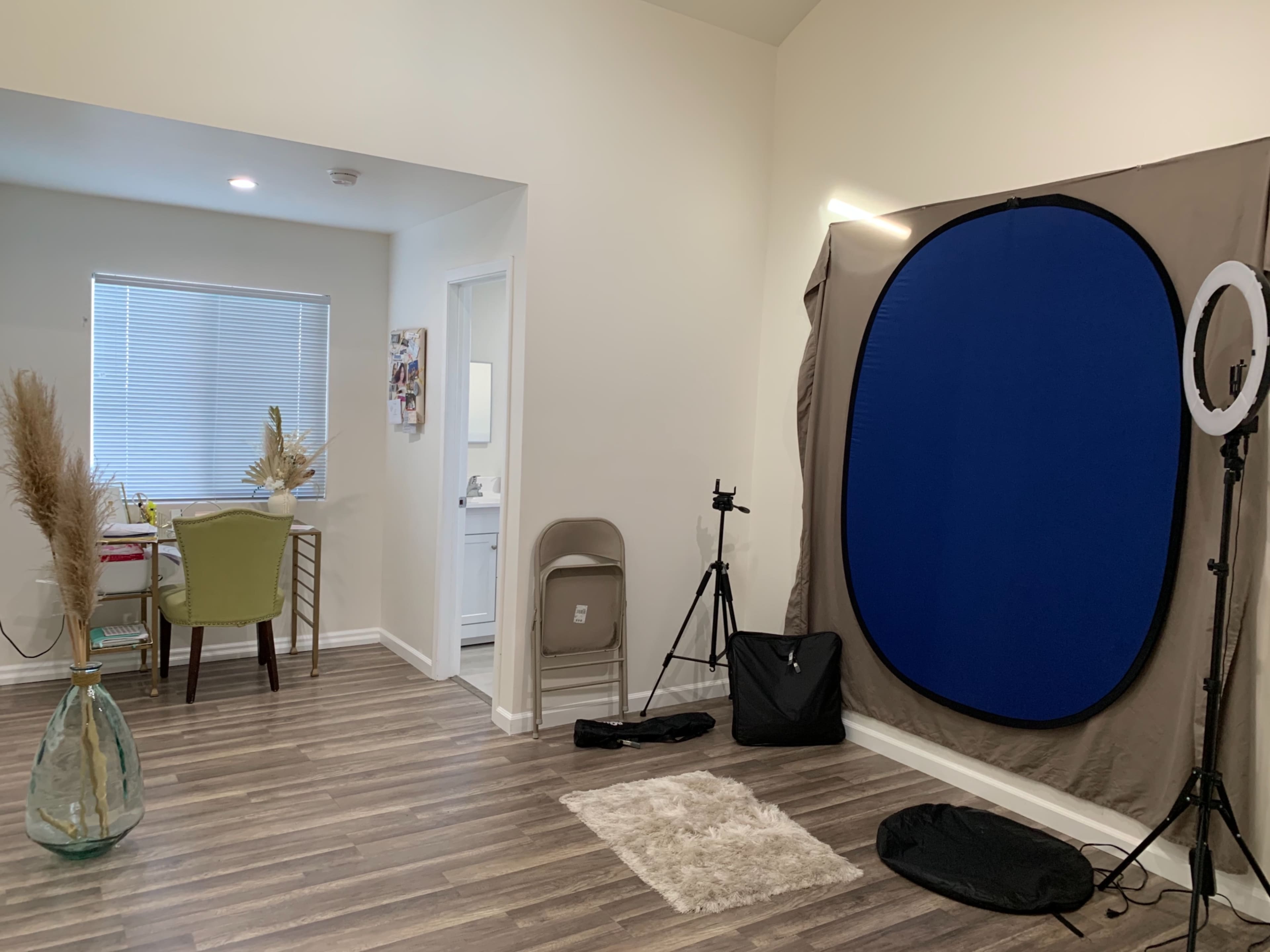 The image shows a well-lit room featuring a small table and chair, a photography backdrop, a ring light, and a decorative vase in the corner.