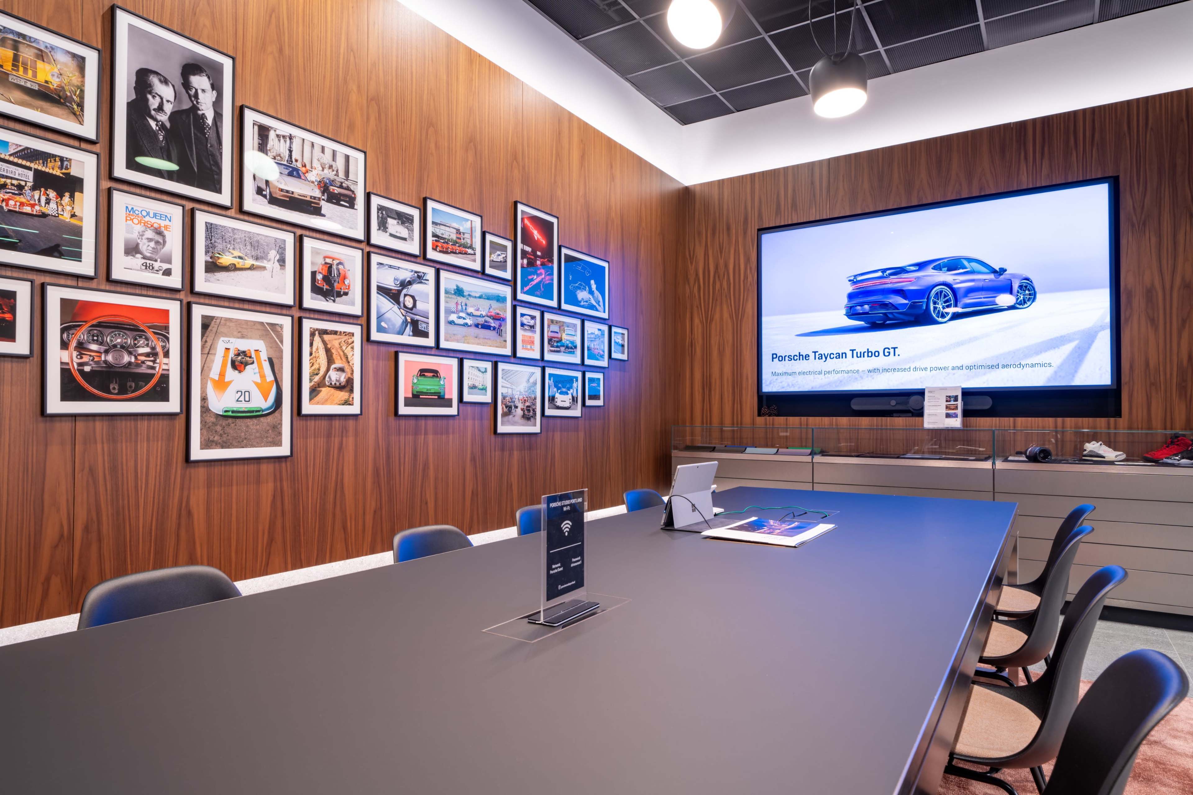 A modern conference room features a sleek black table, a large screen displaying a Porsche Taycan Turbo GT, and an array of framed car-themed photographs on the wooden walls.