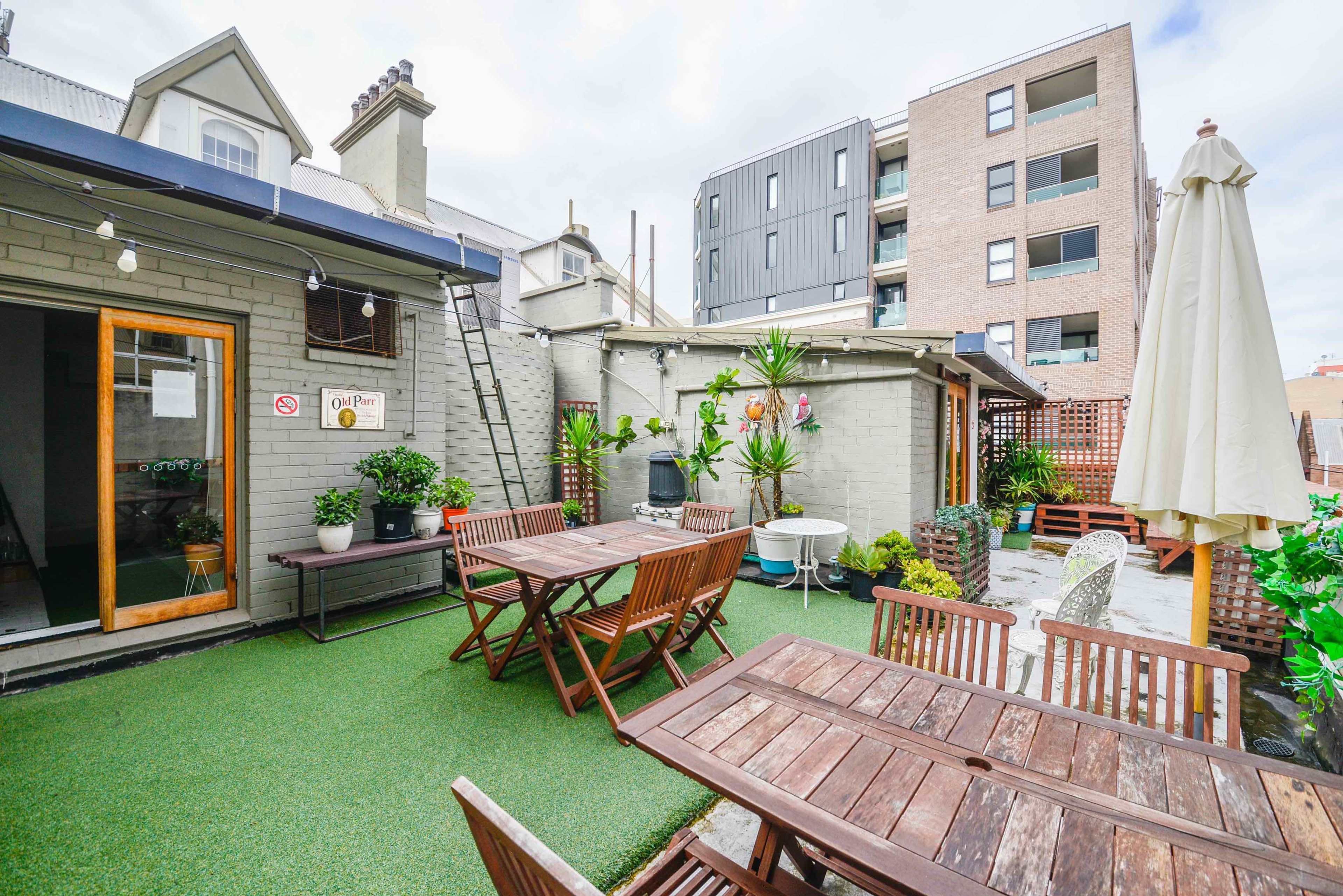 An outdoor patio area with wooden tables and chairs, surrounded by plants and modern buildings in the background.
