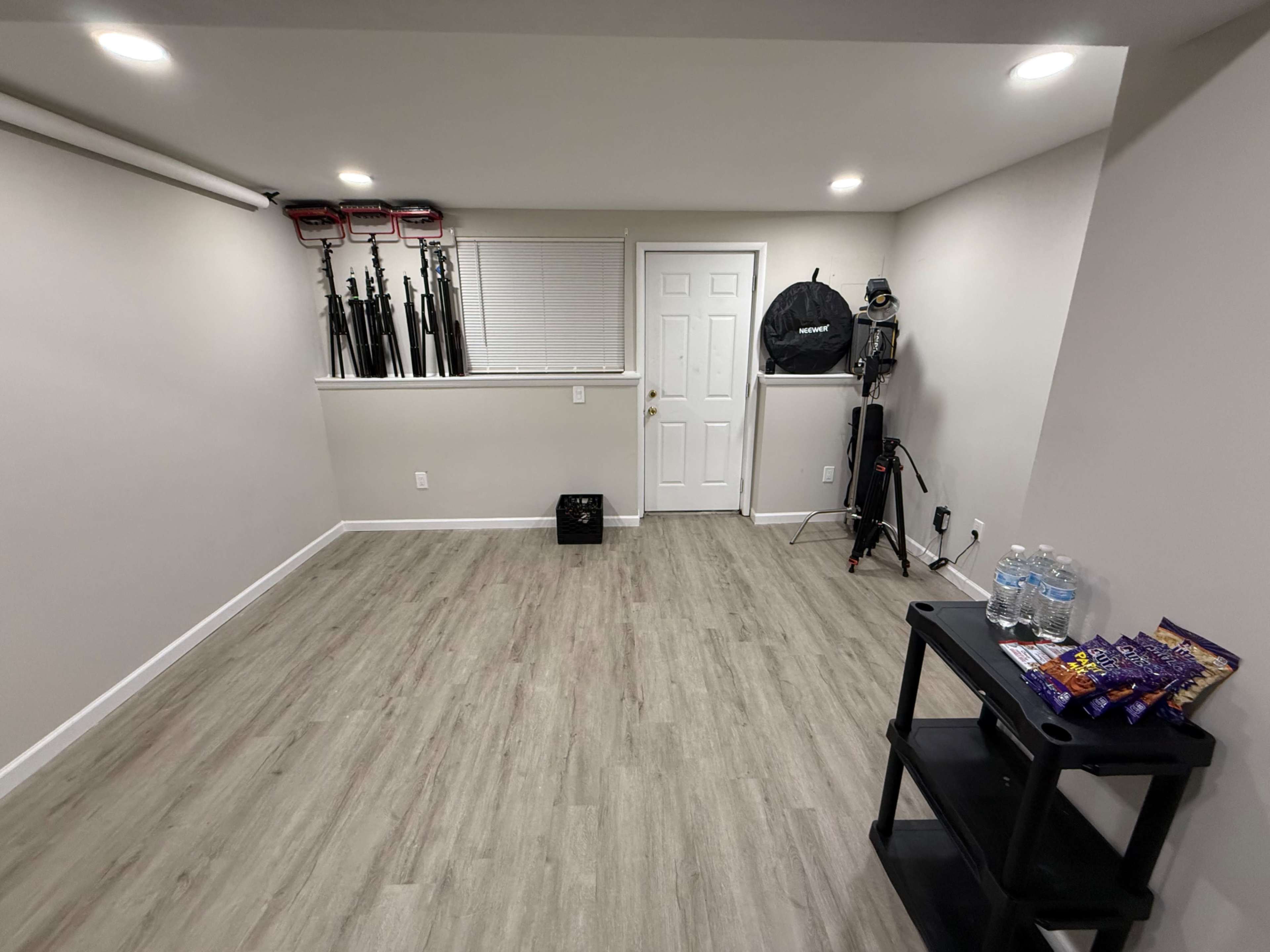 The image shows an empty room with light-colored walls, wooden flooring, and some equipment and water bottles against one wall.
