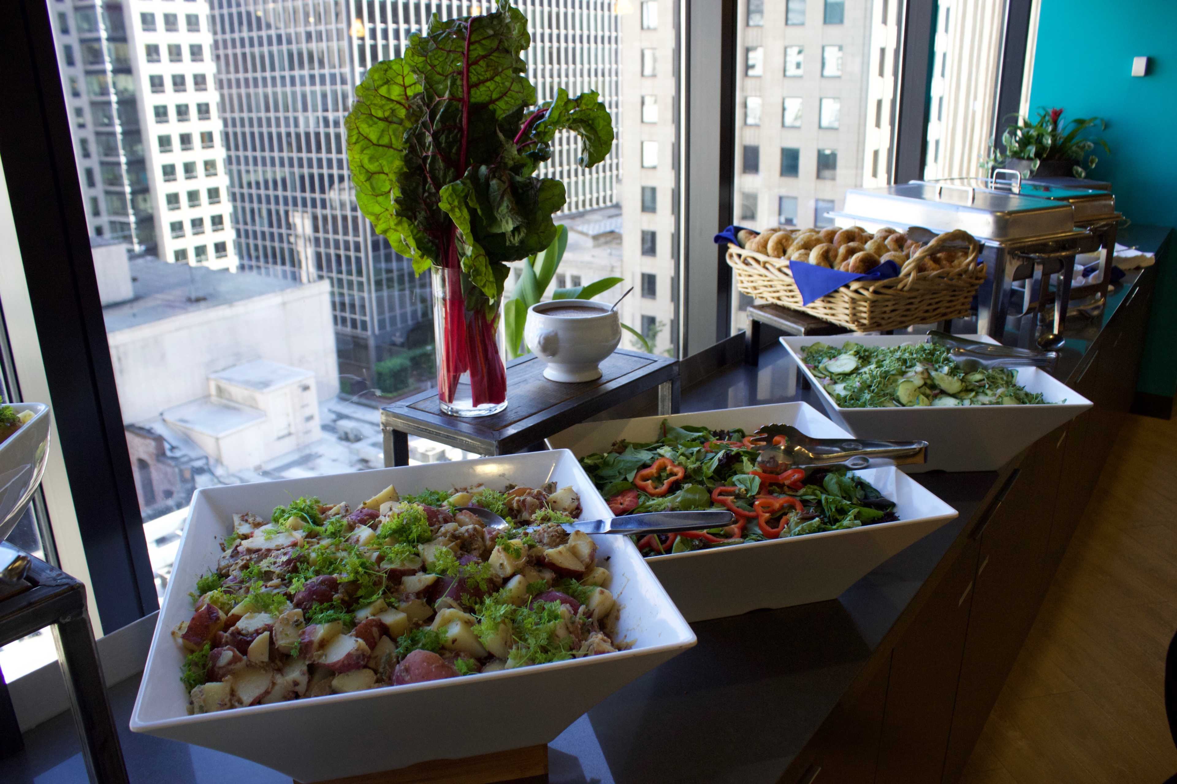 A buffet display features bowls of salad, roasted potatoes, and fresh vegetables near large windows overlooking a cityscape.