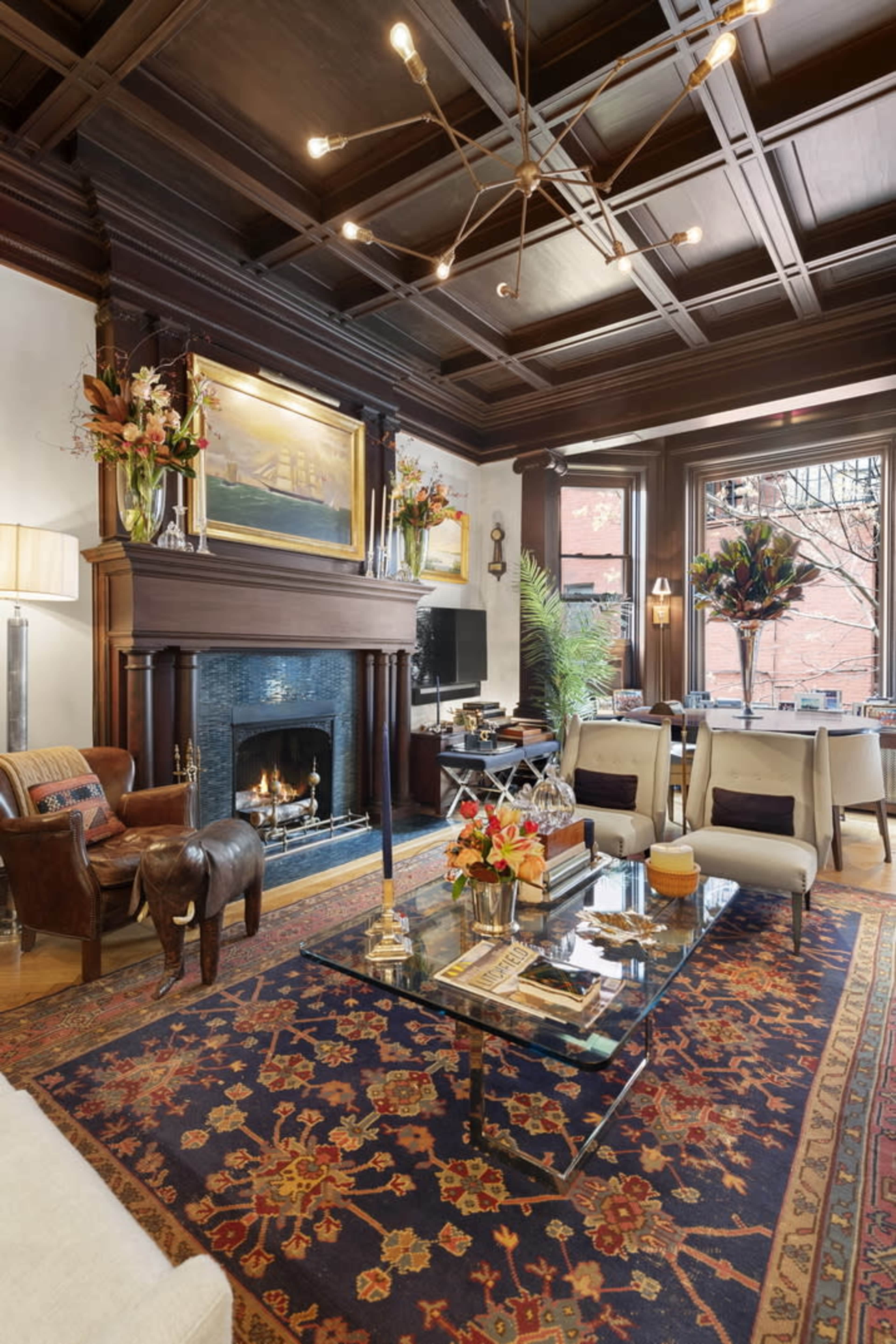 A living room with a coffered ceiling, a fireplace surrounded by a dark wooden mantel, a glass coffee table, and large windows allowing natural light to flood the space.
