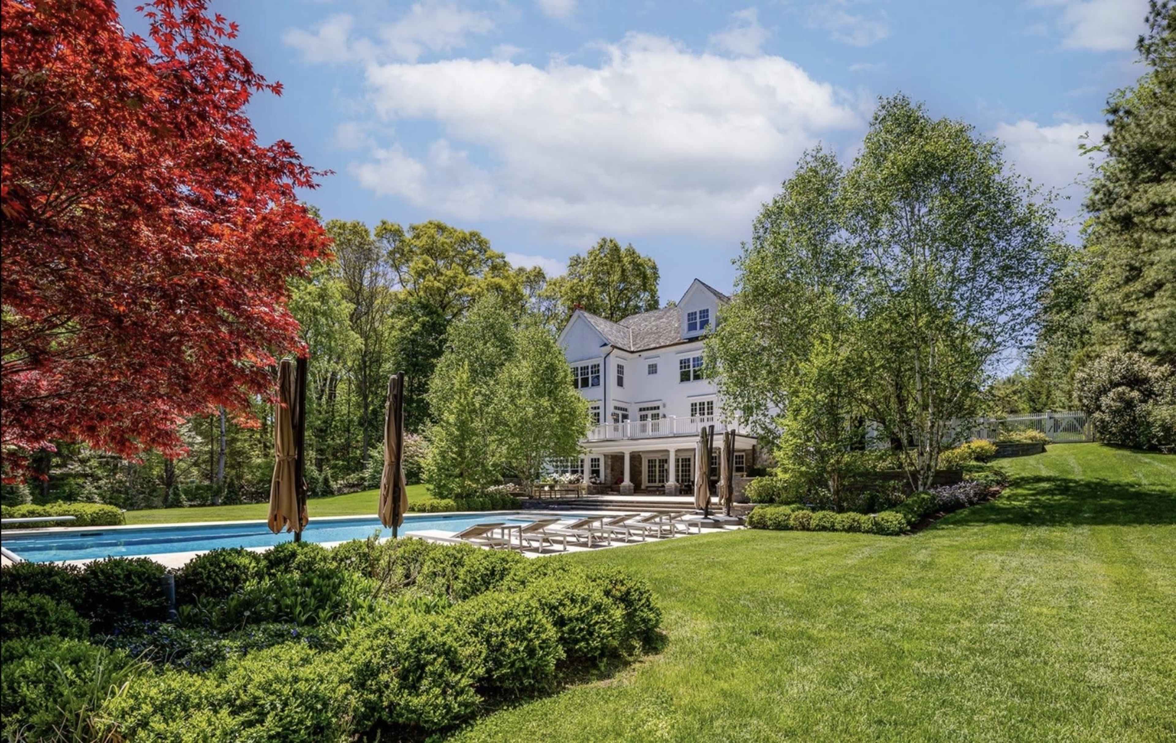 A large white house with a pool is surrounded by well-maintained landscaping and vibrant trees.
