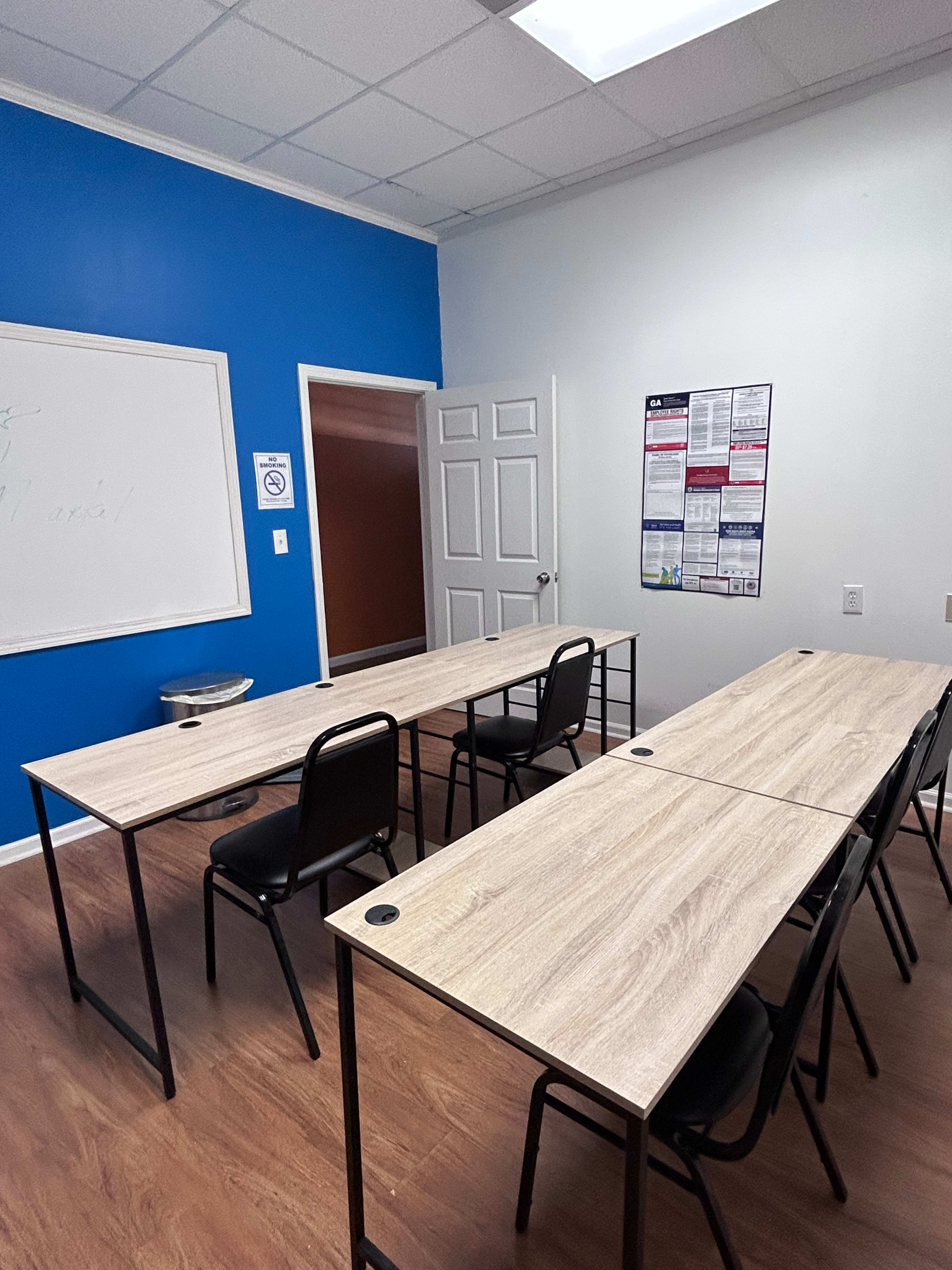 The image shows a classroom with two long tables arranged in a U-shape, surrounded by black chairs, against a blue wall.