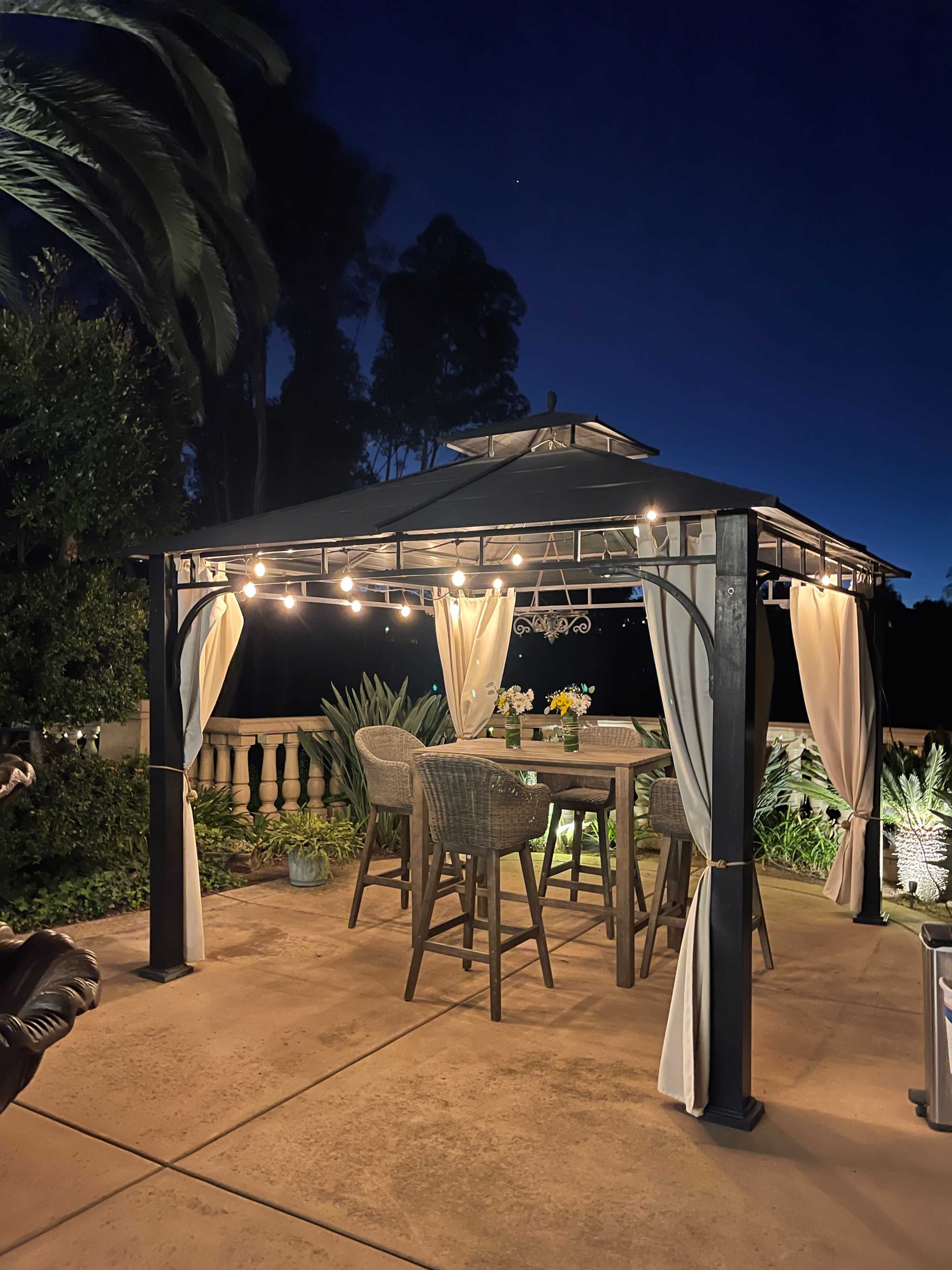 A gazebo with string lights and hanging curtains stands in a patio area at night, surrounded by plants and a decorative railing.