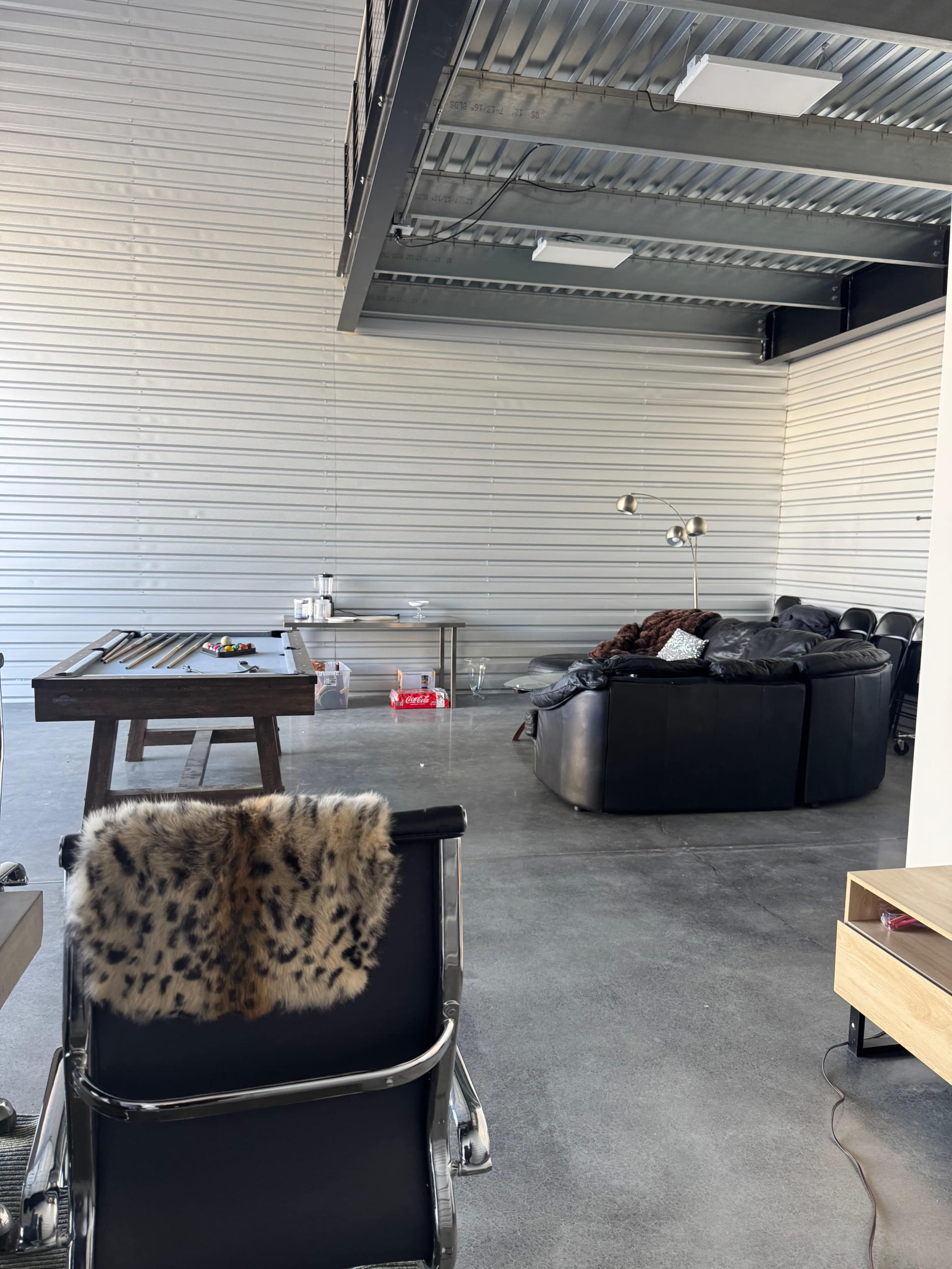 The image shows an industrial-style interior with a seating area featuring a black couch, a wooden table with tools, and a metal overhead structure.