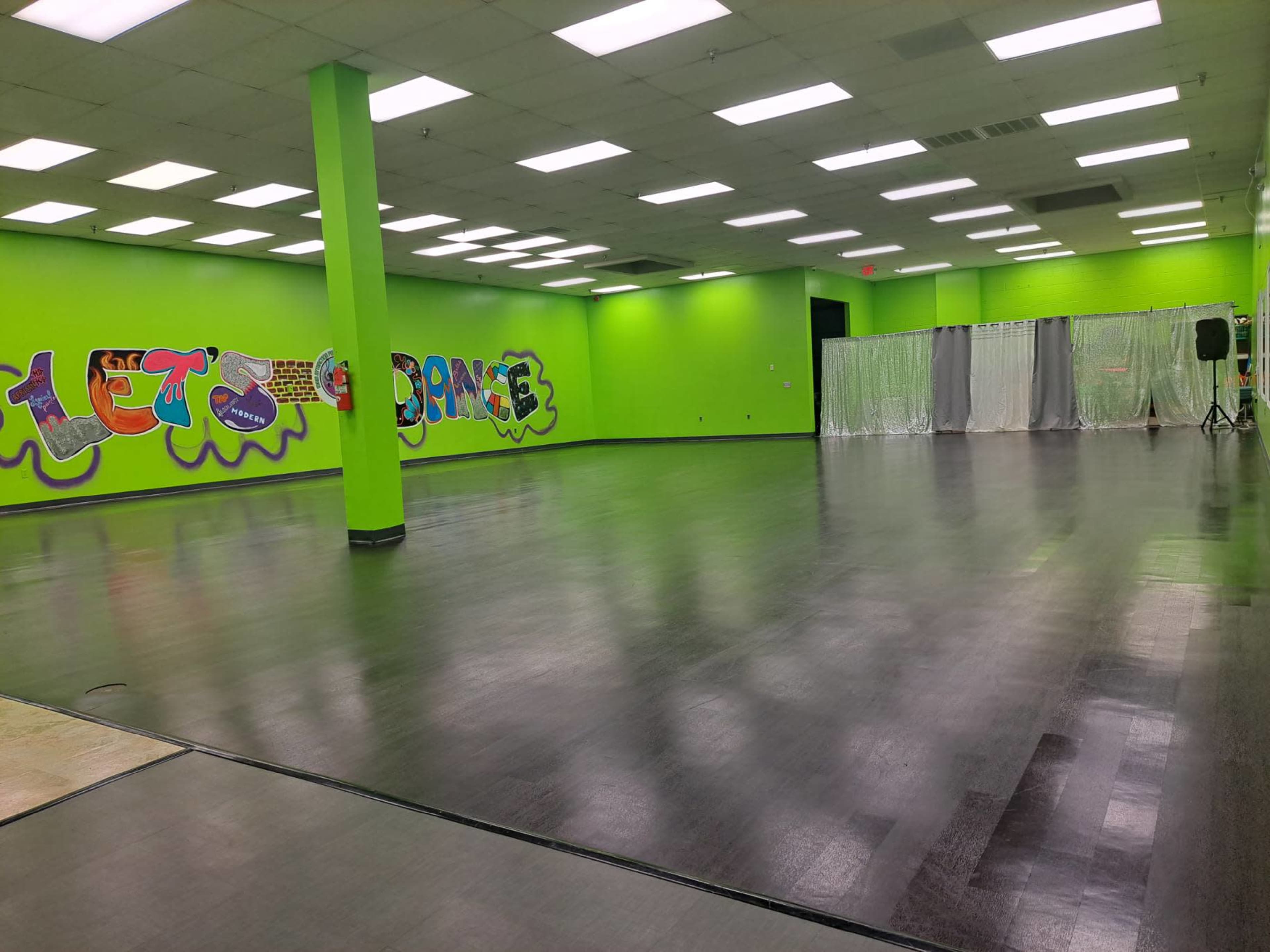 A spacious dance studio with vibrant green walls, a polished black floor, and bright overhead lights.