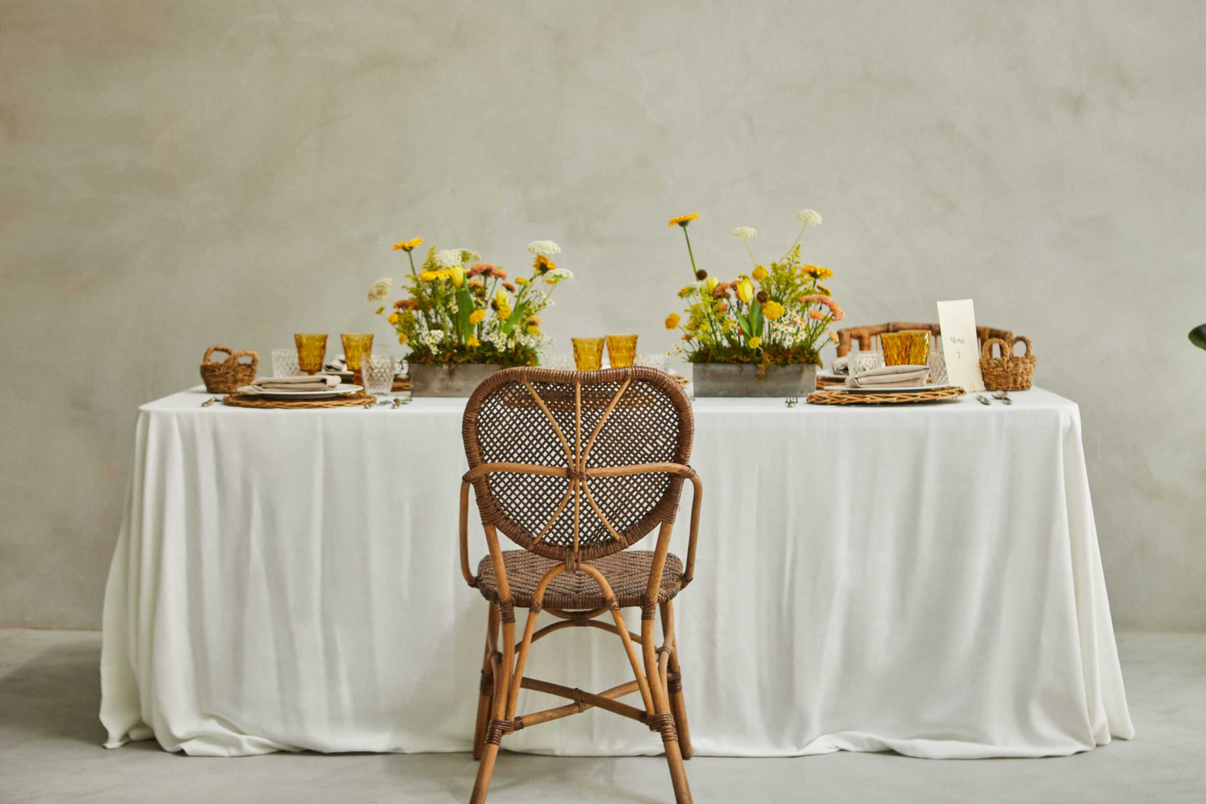 A rustic dining table is set with floral centerpieces, glassware, and woven placemats, accompanied by a single wicker chair facing the table.
