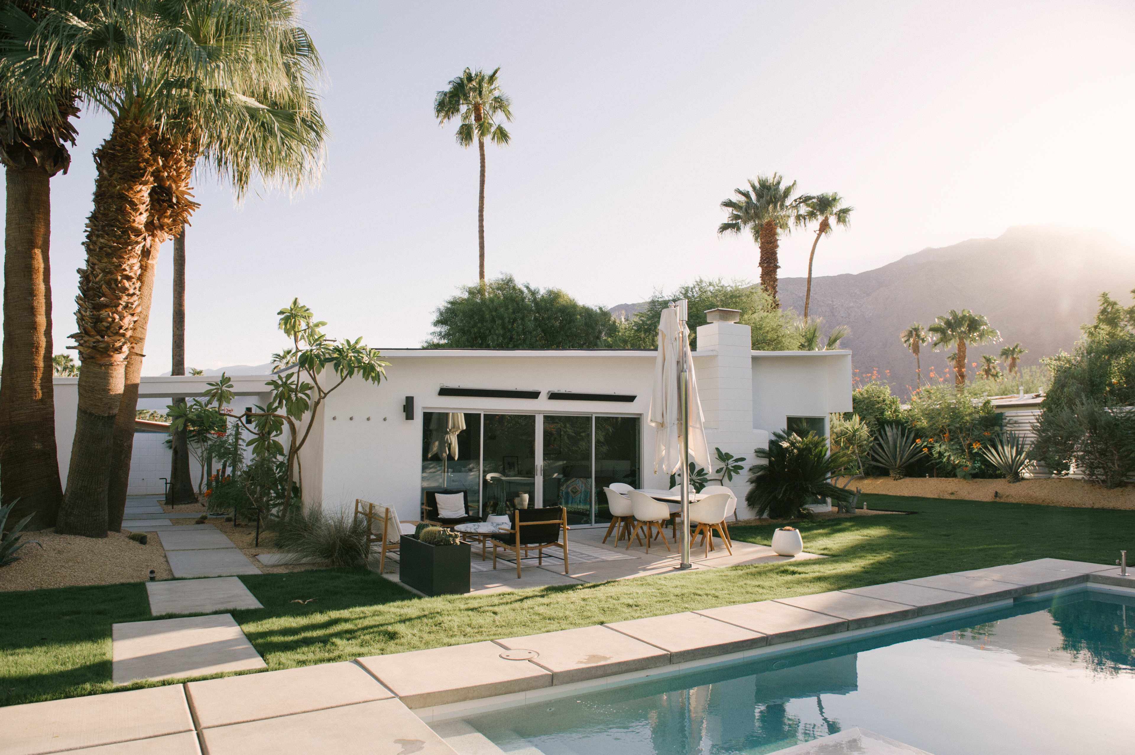 A modern white house with large windows is surrounded by palm trees and a swimming pool, set against a mountainous backdrop.